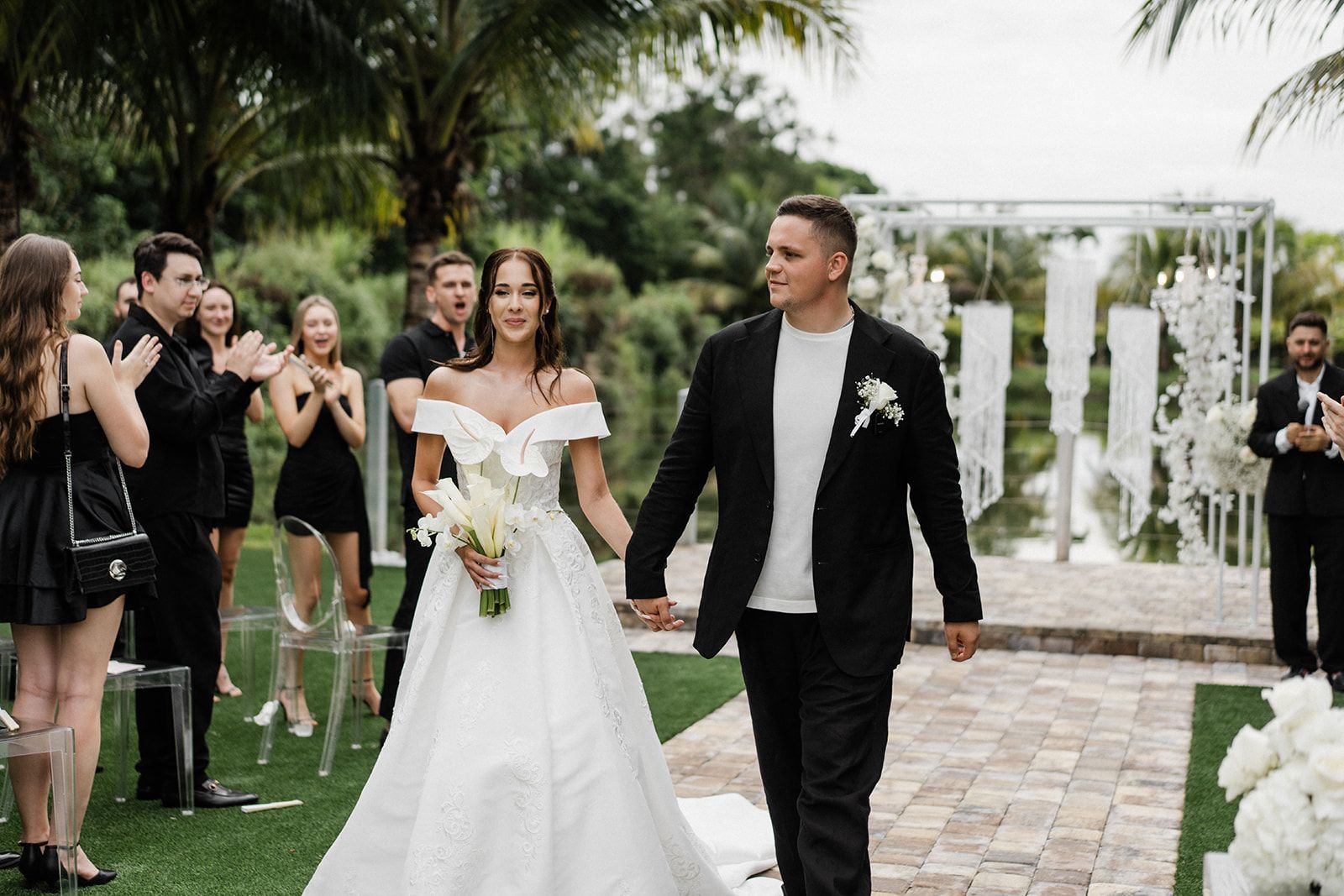 bride and groom hold hands and walk out of the ceremony with minimal white and airy wedding arch behind them 