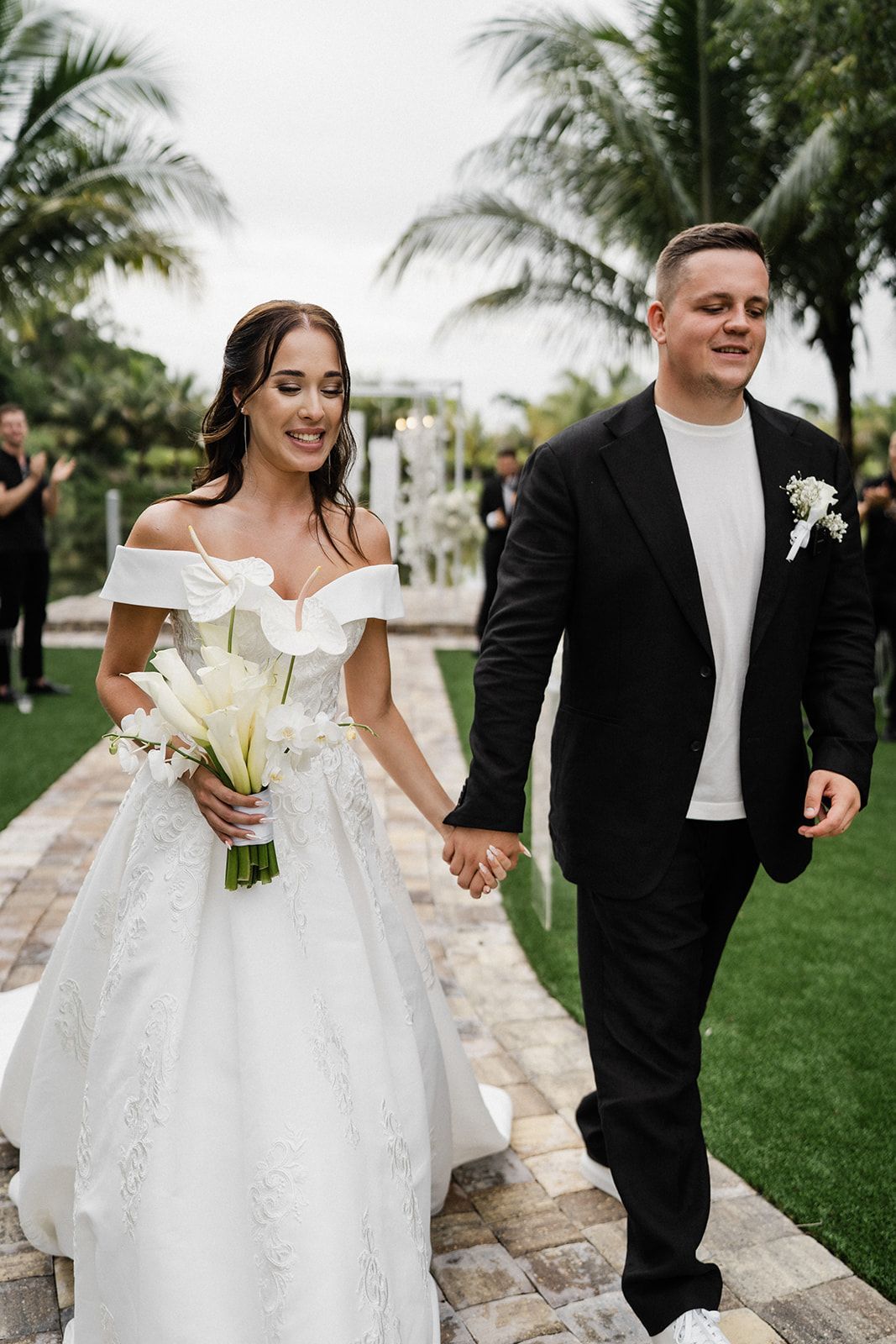 Bride and groom holding hands, smiling, walking down aisle. White dress, black suit, outdoor wedding.