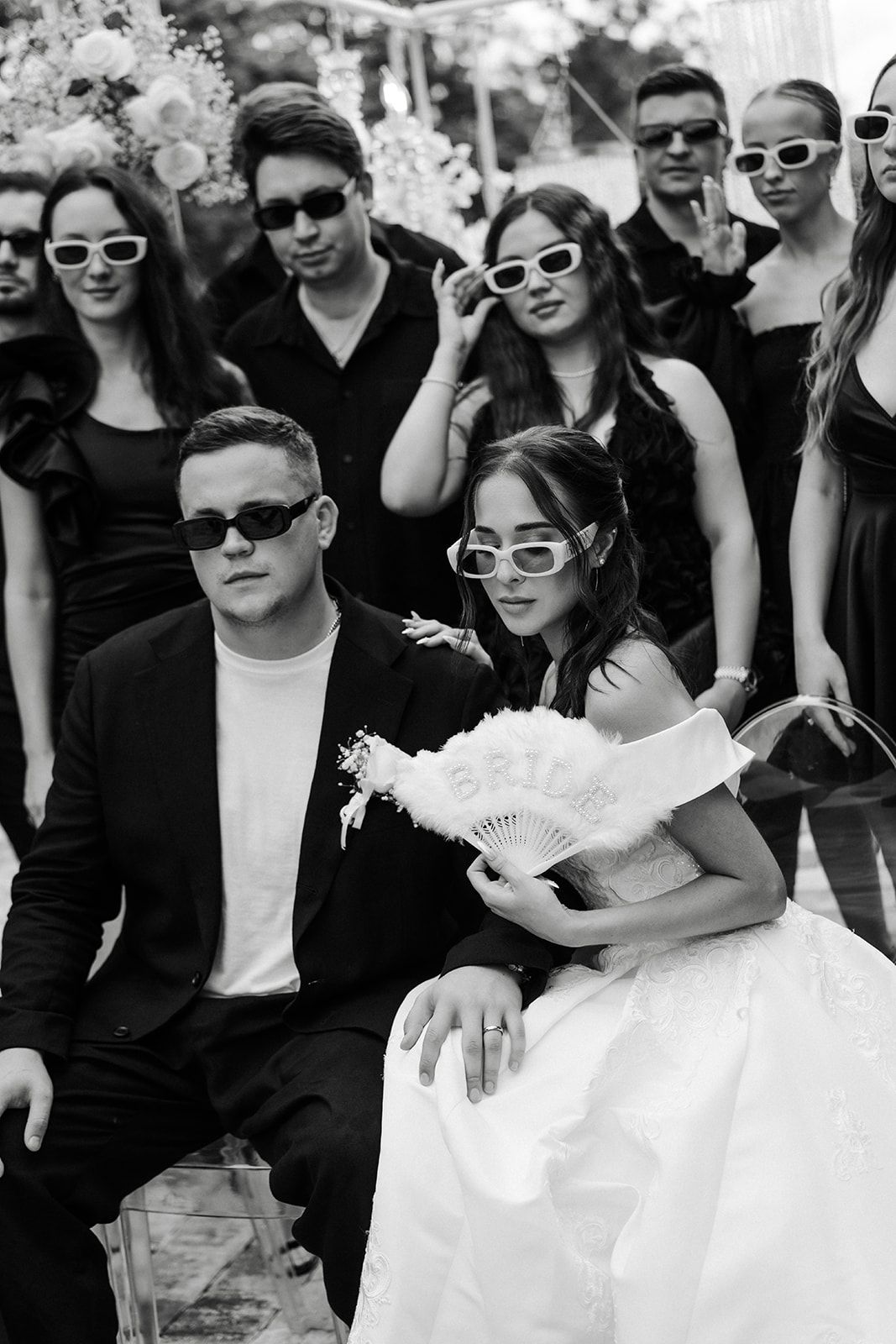 Group in black outfits and white sunglasses with a bride in a white dress, posing outdoors.