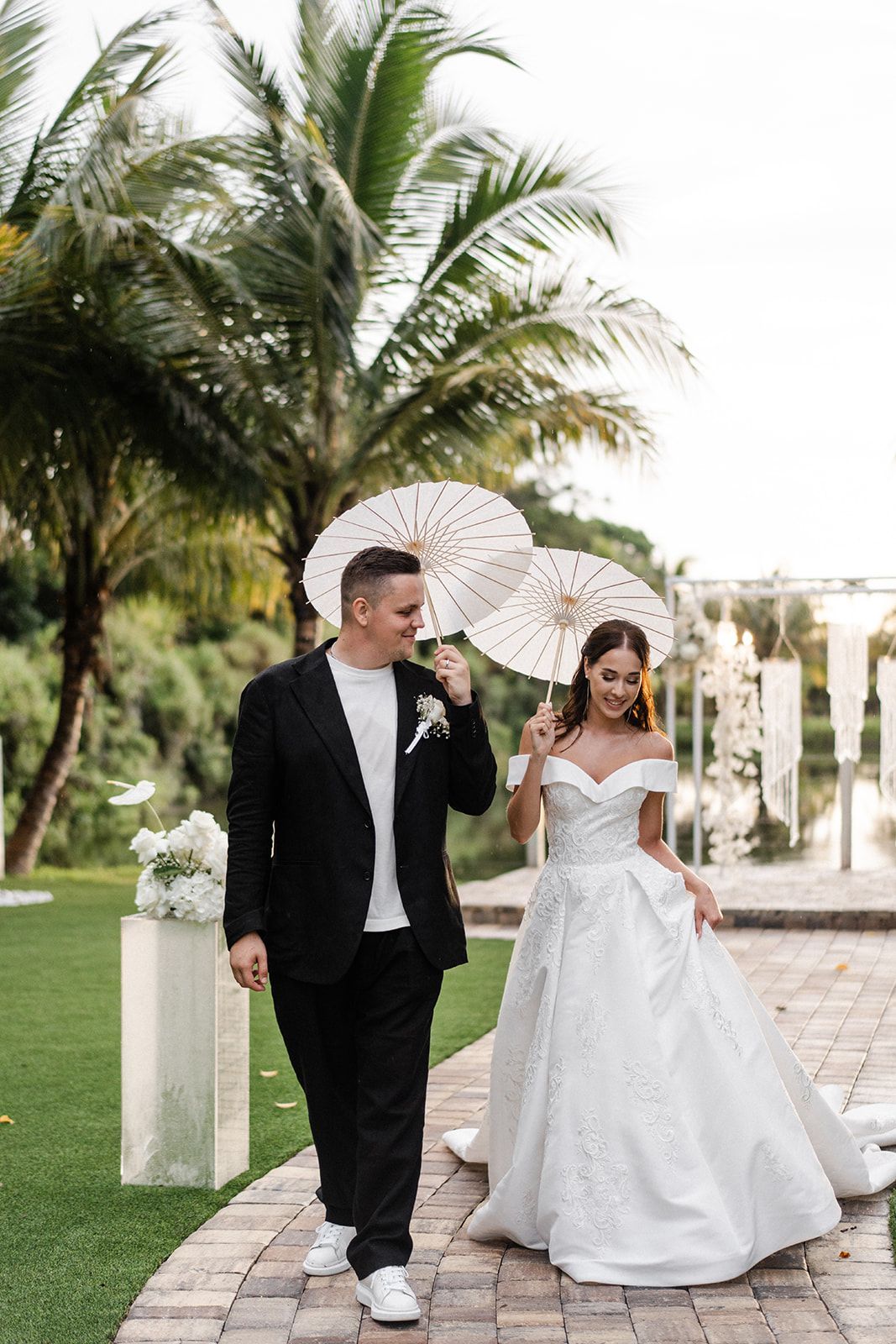 Bride and groom walk, holding parasols, on a brick path, near a body of water and palm trees.