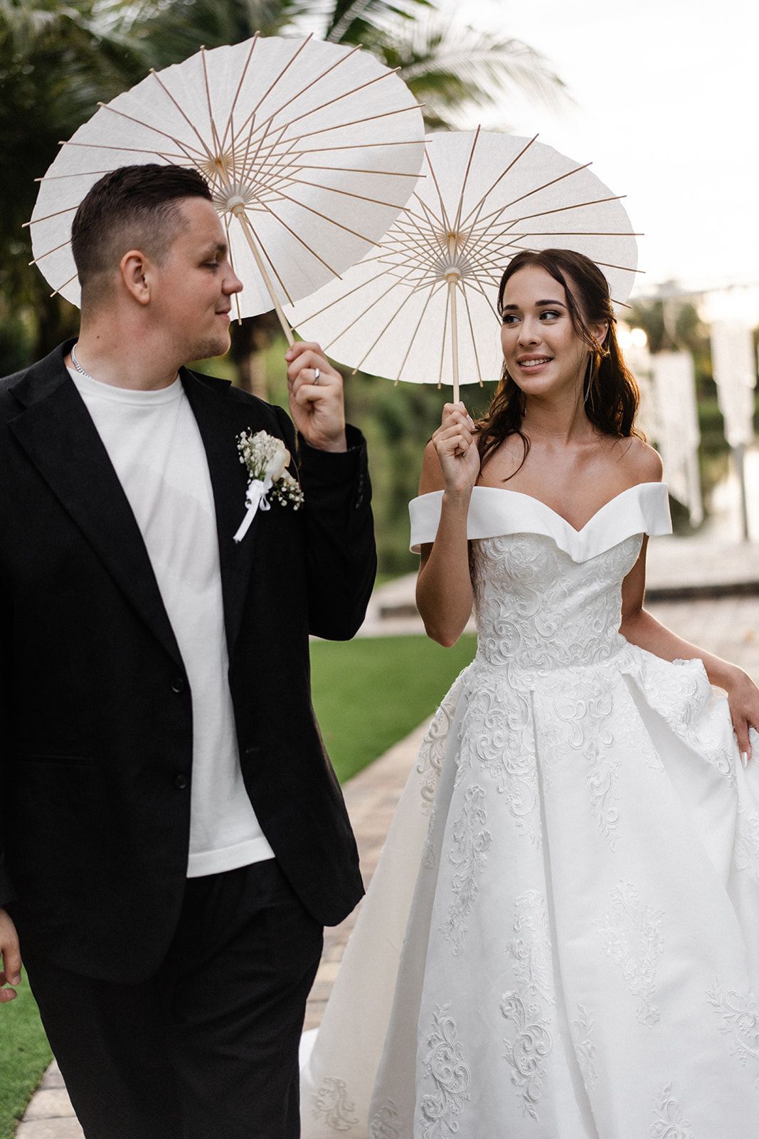 Newlyweds walking outdoors, holding white parasols. Bride in white gown smiles at groom in black suit.