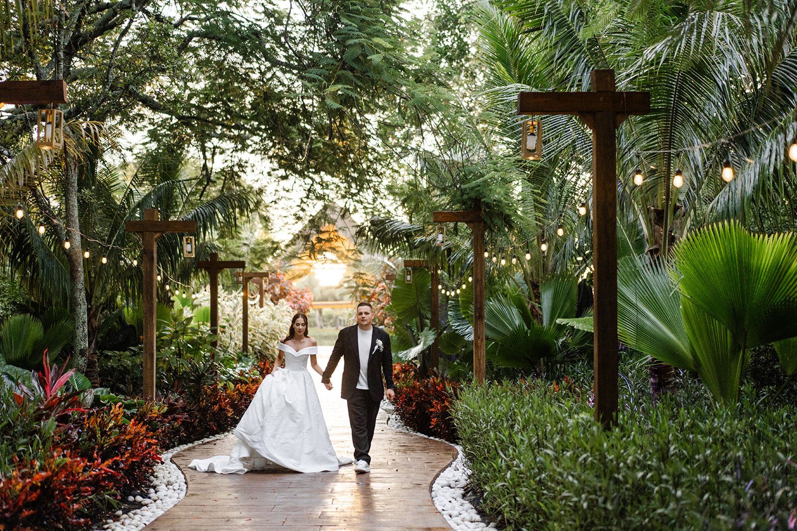 Bride and groom holding hands, walking along a pathway lined with lights, surrounded by lush greenery, at a wedding.