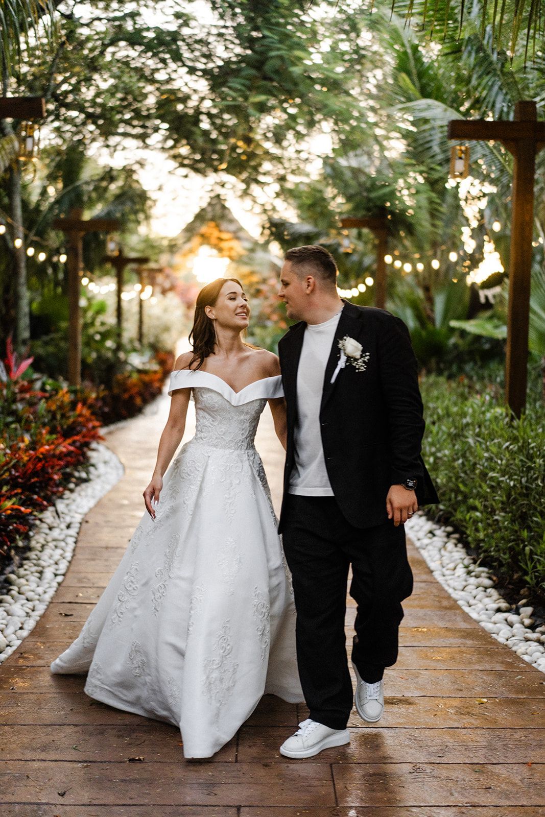 Couple walking hand-in-hand on path, woman in wedding gown, man in suit, outdoor setting with plants and lights.
