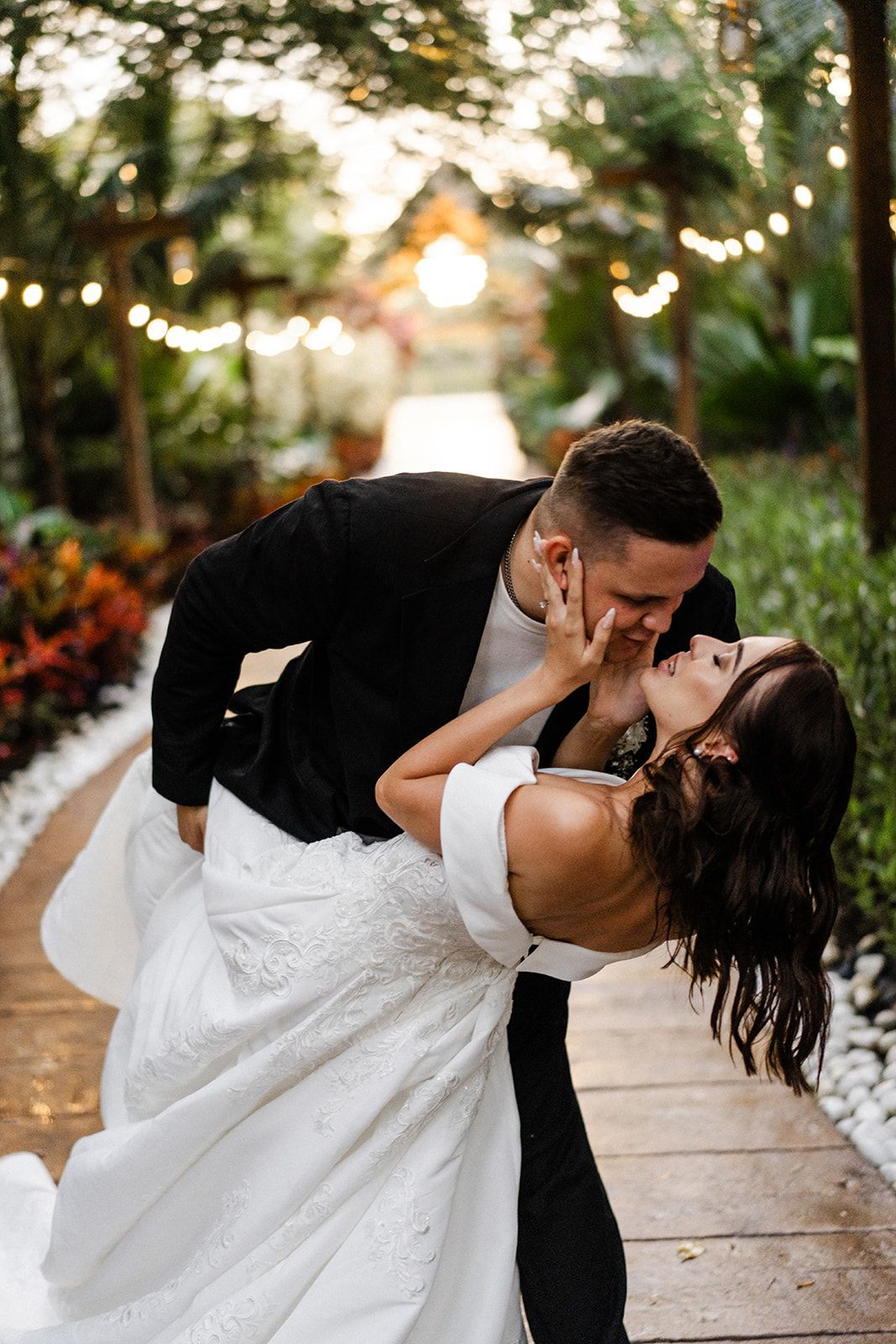 Couple dancing and kissing outdoors. Bride in white gown, groom in black blazer, wooden path with string lights.