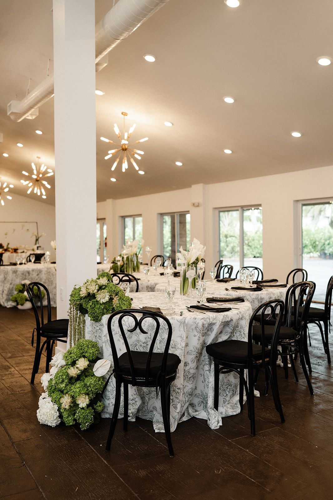 Elegant event space with round tables, black chairs, and white lace tablecloths; green and white floral arrangements.