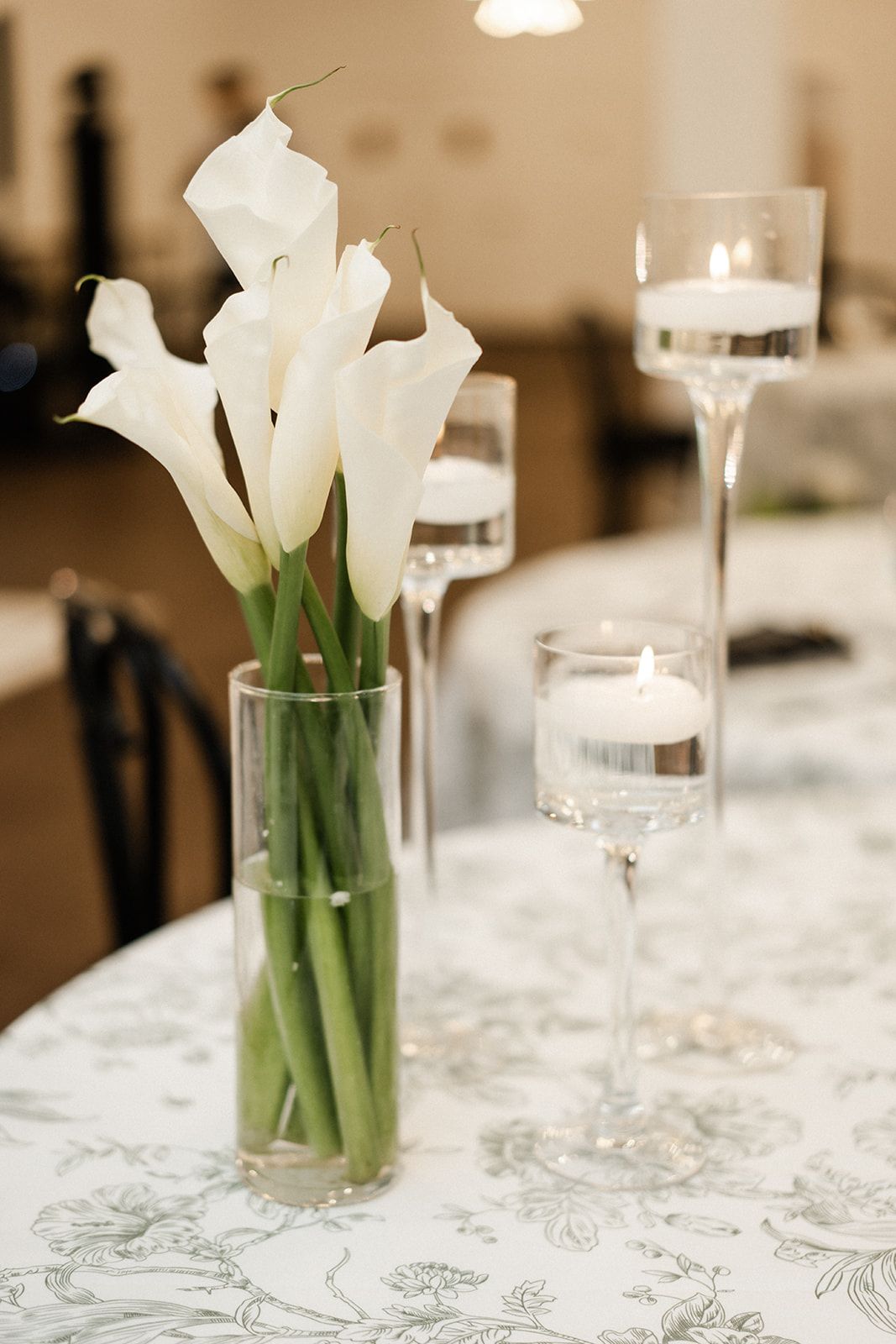 White calla lilies in a vase, with floating candle centerpieces on a floral tablecloth.