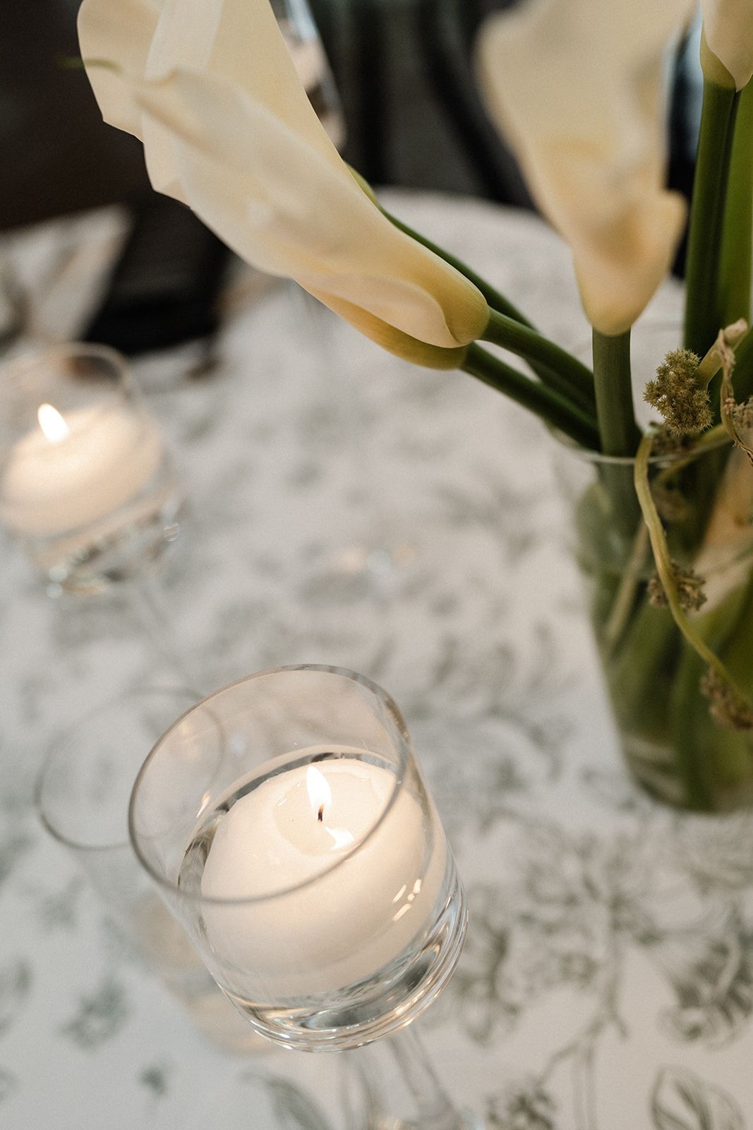White calla lilies in vase on floral tablecloth, with two lit votive candles.