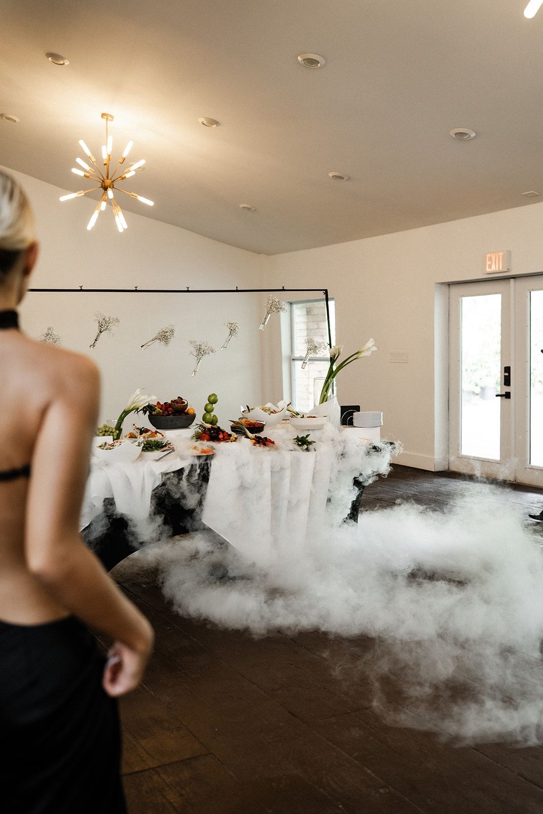 Woman in black attire observes table set for a meal with fog. White walls, gold chandelier, and flowers.