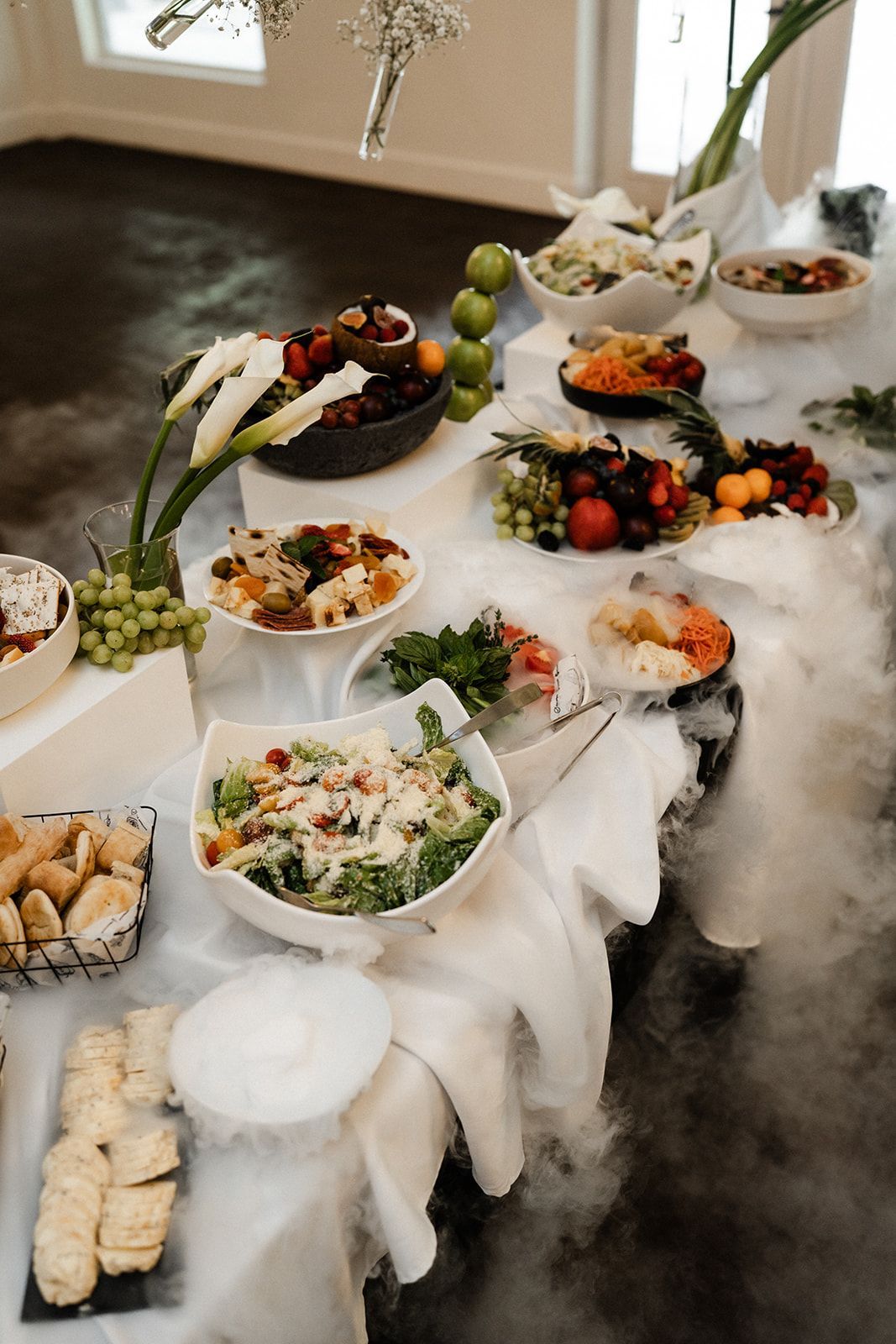 Buffet table with various salads and fruits, surrounded by dry ice, creating a smoky effect.