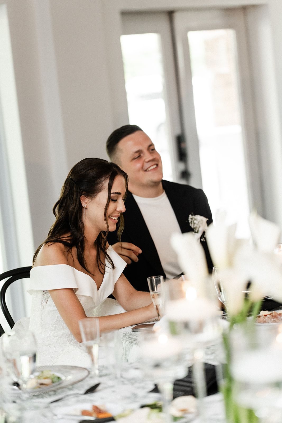 Bride and groom at a wedding reception, smiling, seated at a table. White dress, black suit.