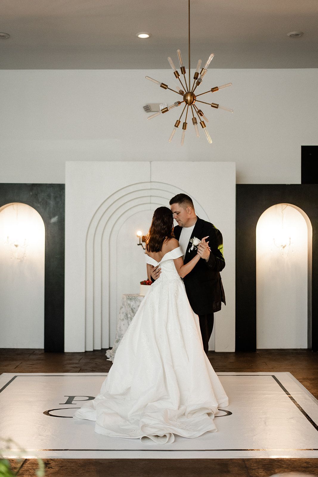 Couple dances at their wedding reception. Bride in white dress, groom in black suit, in front of white backdrop, and light fixtures.
