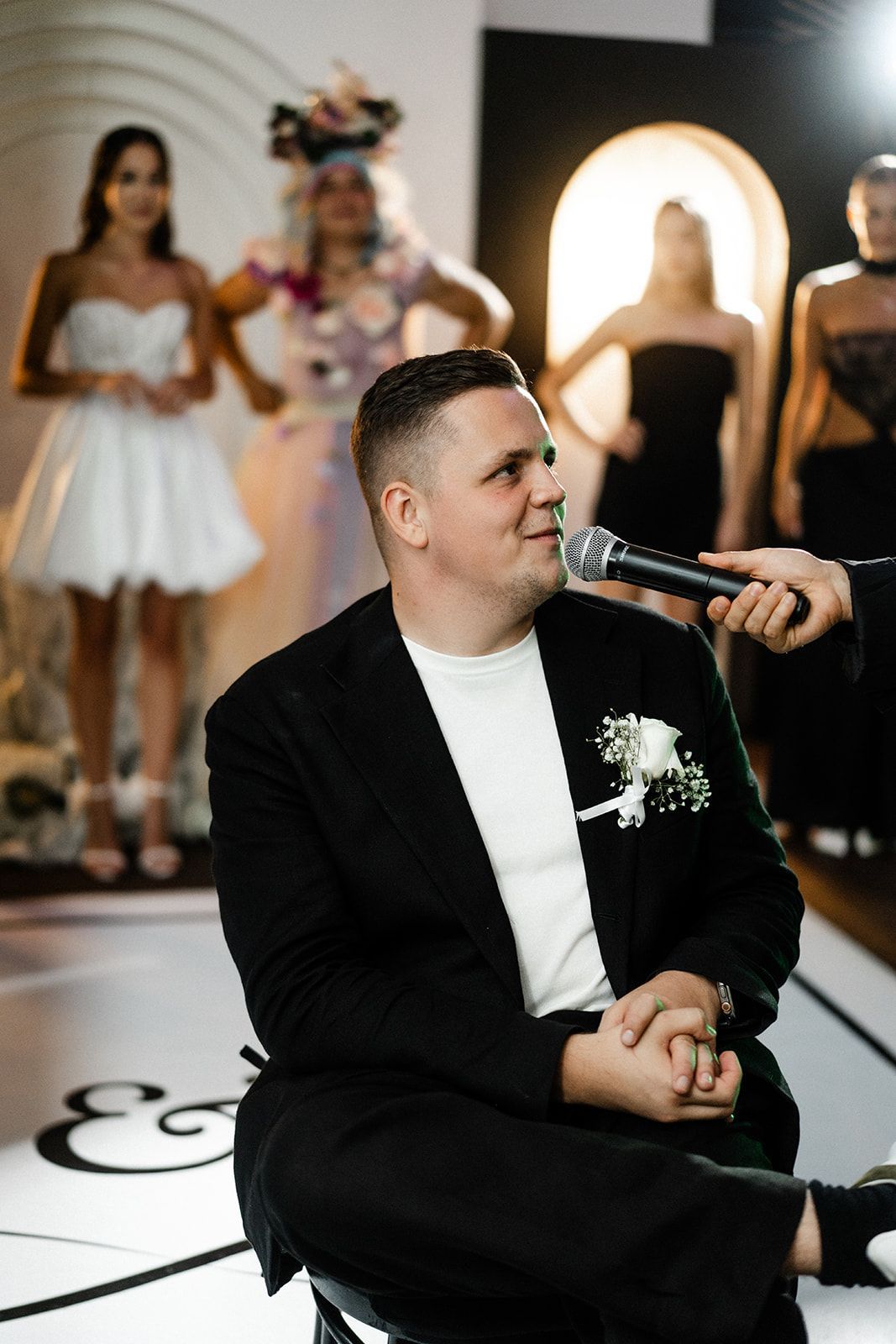 Man in a black suit being interviewed, models in the background.