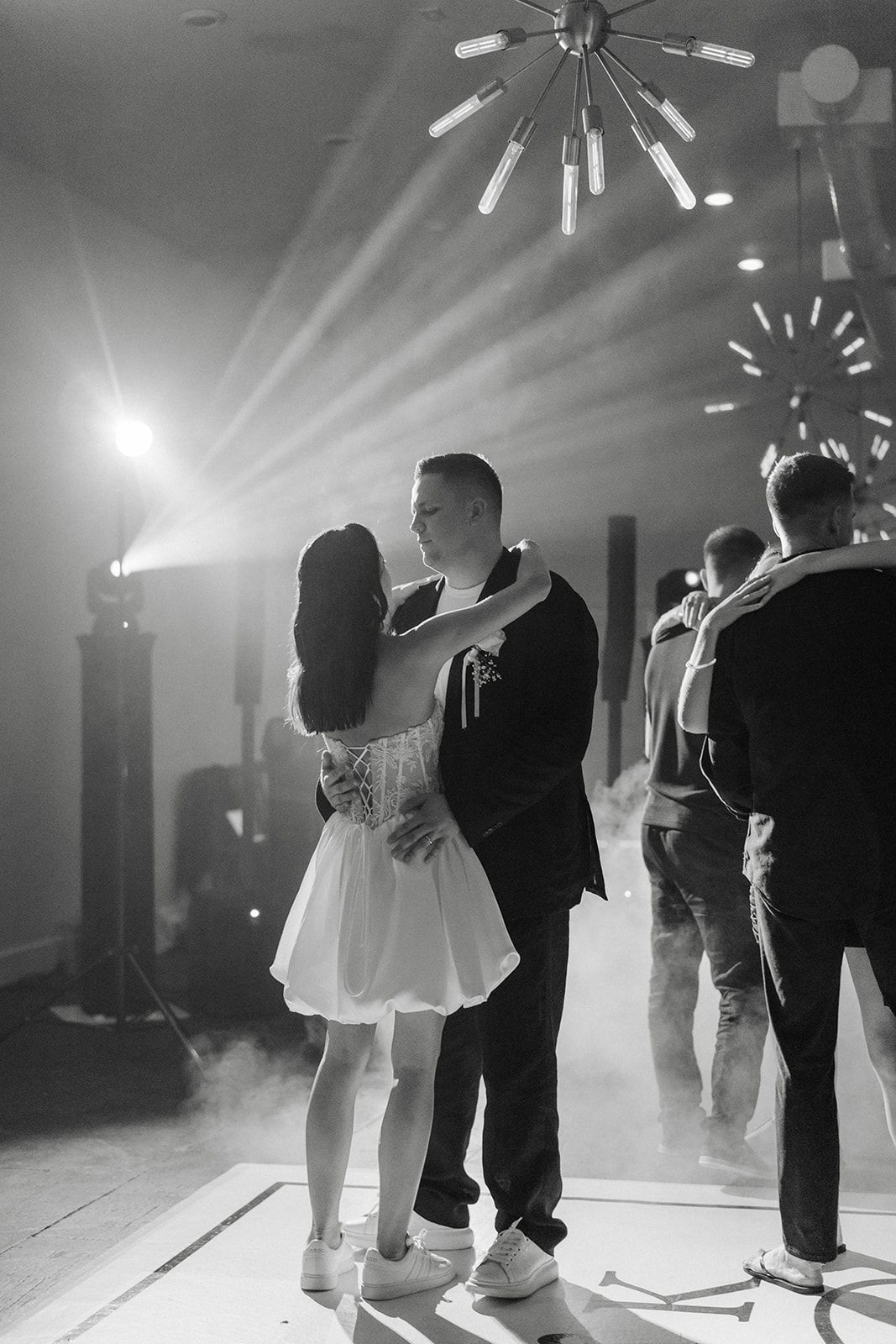 Couple dancing in a spotlight on a dance floor, other guests visible in the background. Black and white.