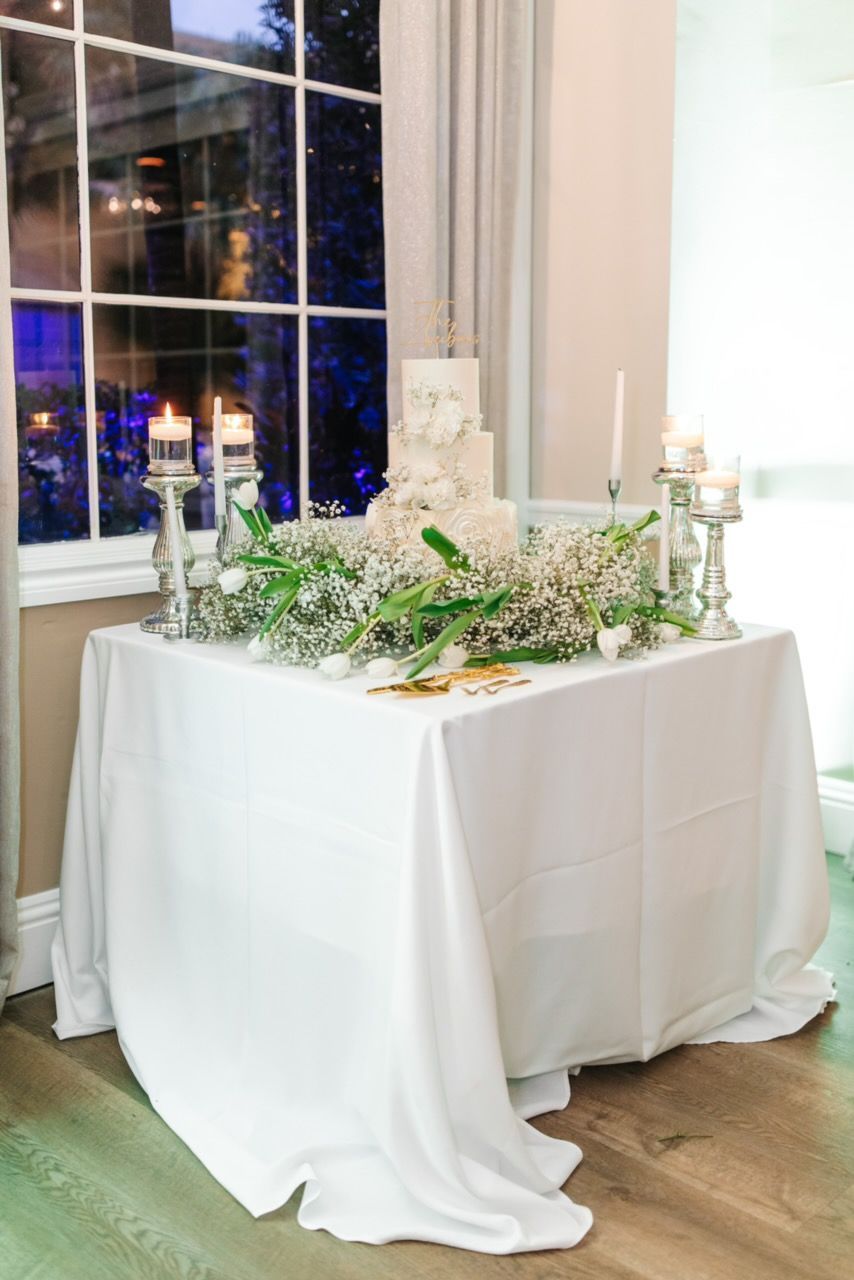 Wedding cake on a white-draped table with candles and flowers; window in the background.