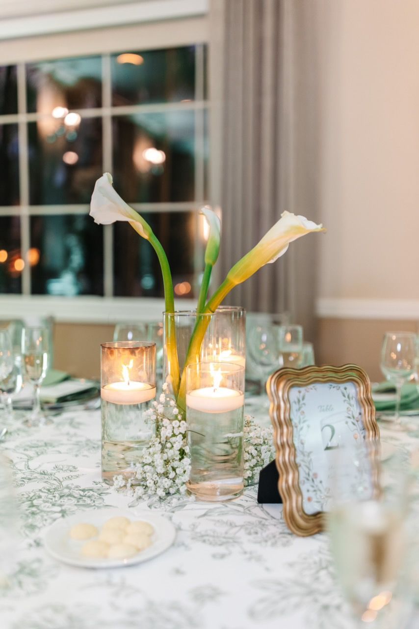 Elegant wedding table centerpiece with calla lilies, floating candles, and framed table number.