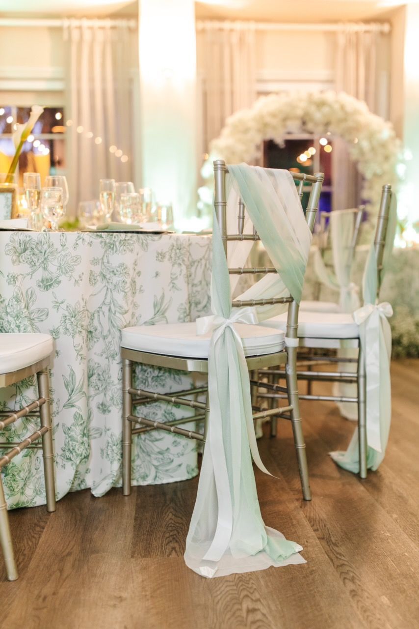 Elegant wedding reception: Chairs draped with light green fabric; floral tablecloth; arch with white flowers.