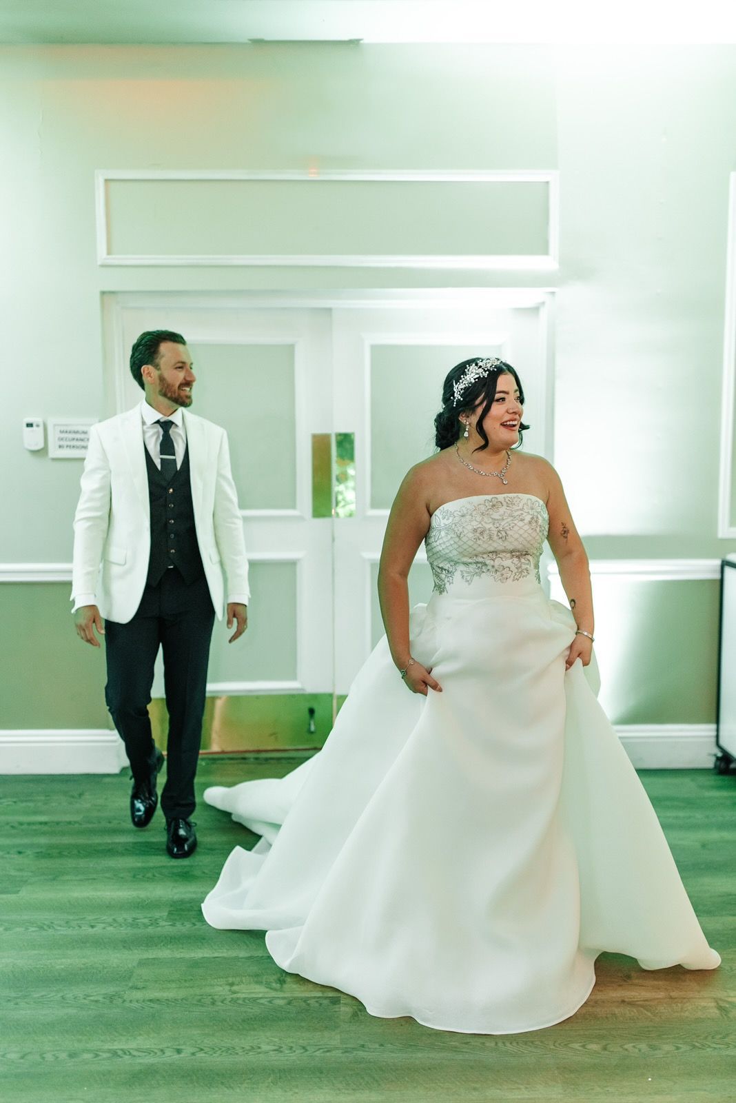 Bride in strapless gown, groom in white jacket walk towards doors.