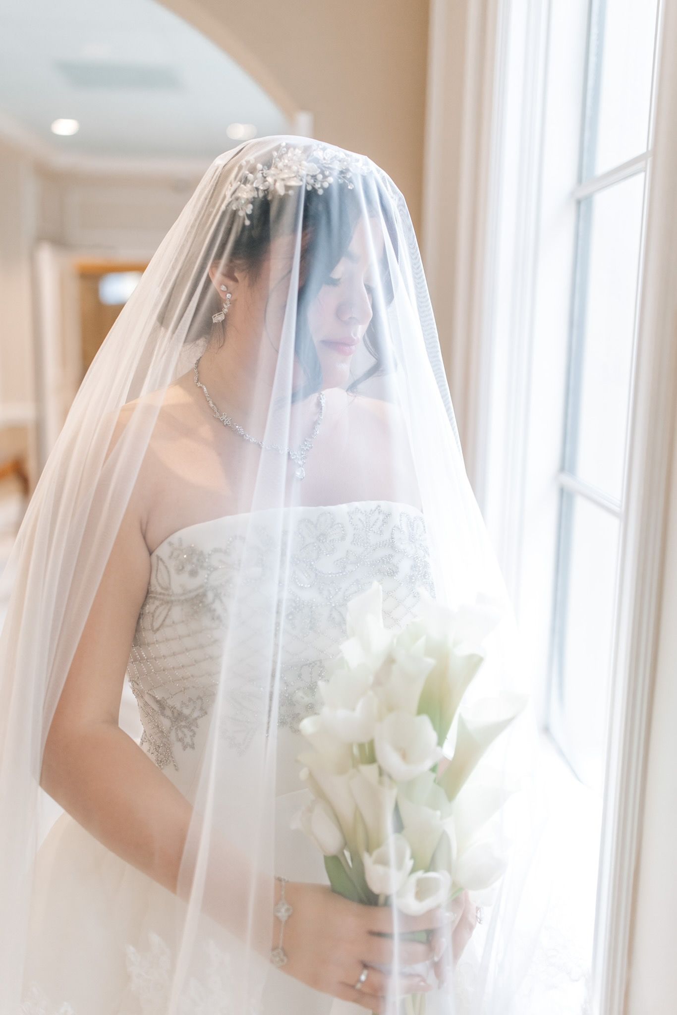 Bride in strapless, beaded gown, veil covering her face, holding white flowers by a window.