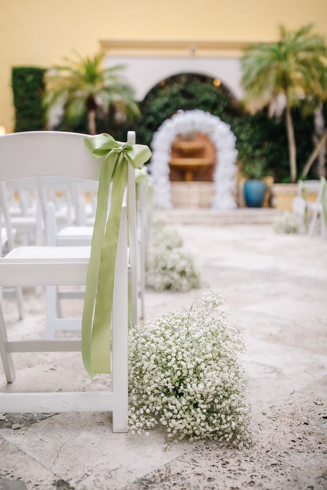 White chairs with green ribbon and baby's breath mark a wedding aisle in a courtyard.