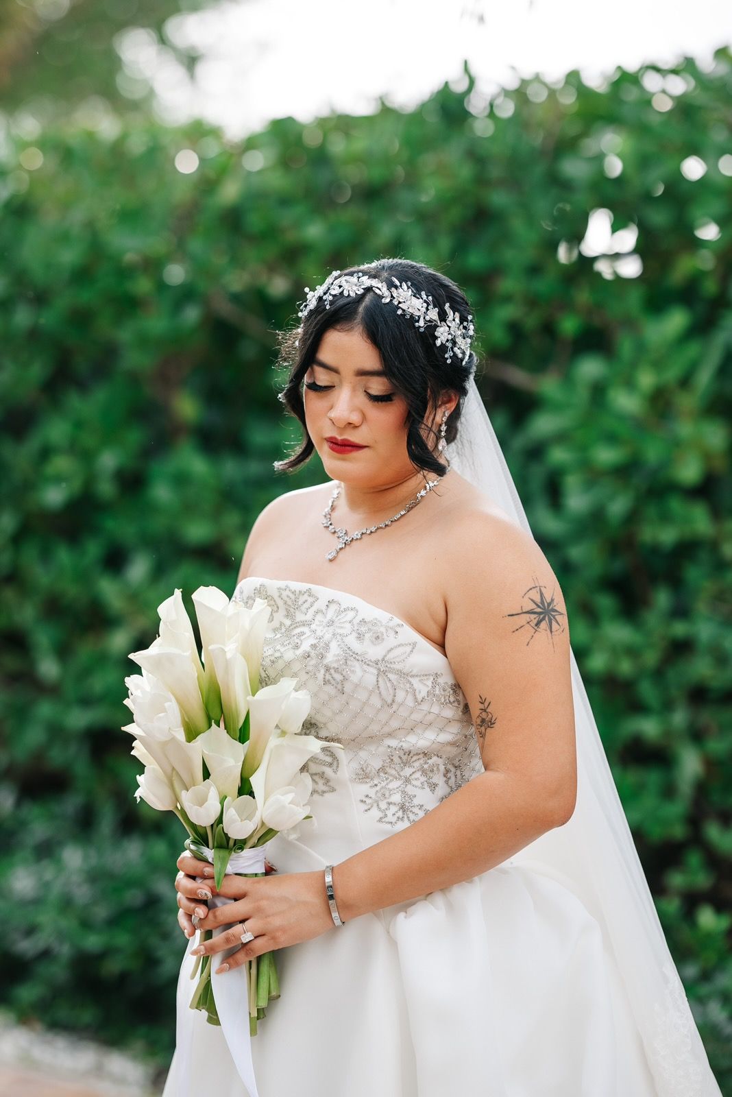 Bride in a strapless gown holds a bouquet of white flowers, looking down. Green foliage background.