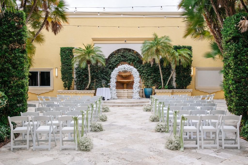 Wedding ceremony setup in an outdoor courtyard. White chairs line the aisle leading to an archway and backdrop of greenery.