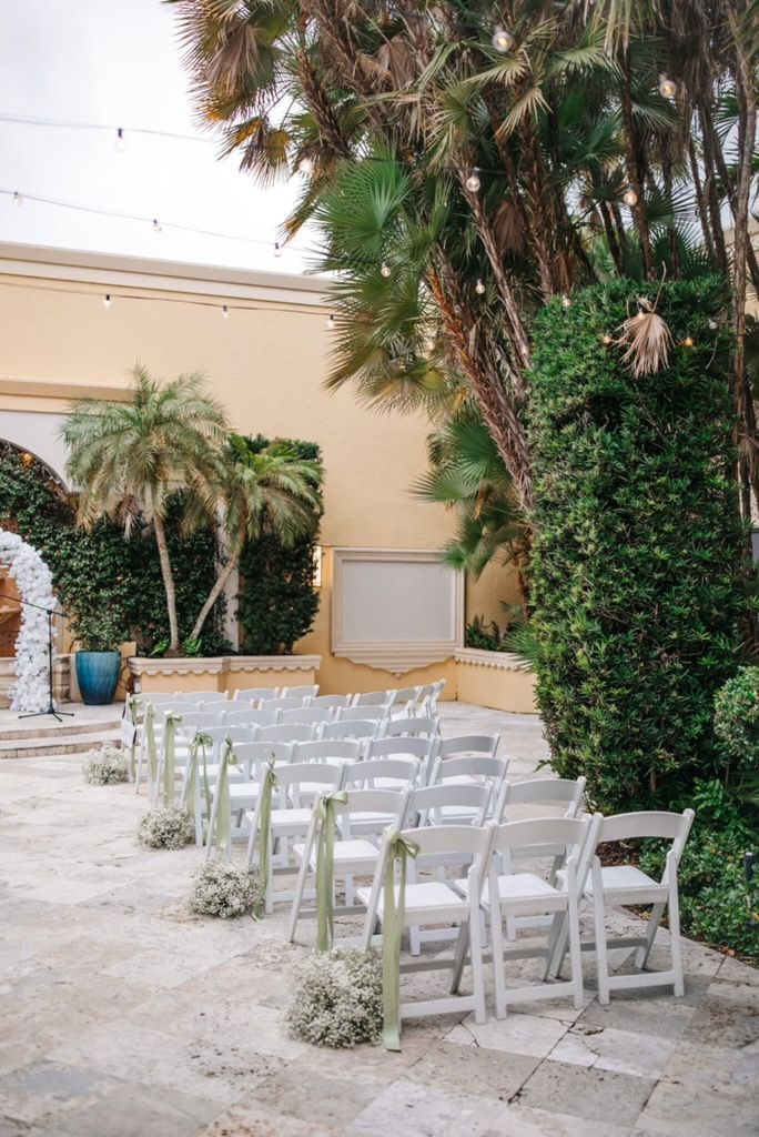 Rows of white chairs with ribbons arranged for an outdoor wedding ceremony, with floral decorations and greenery.