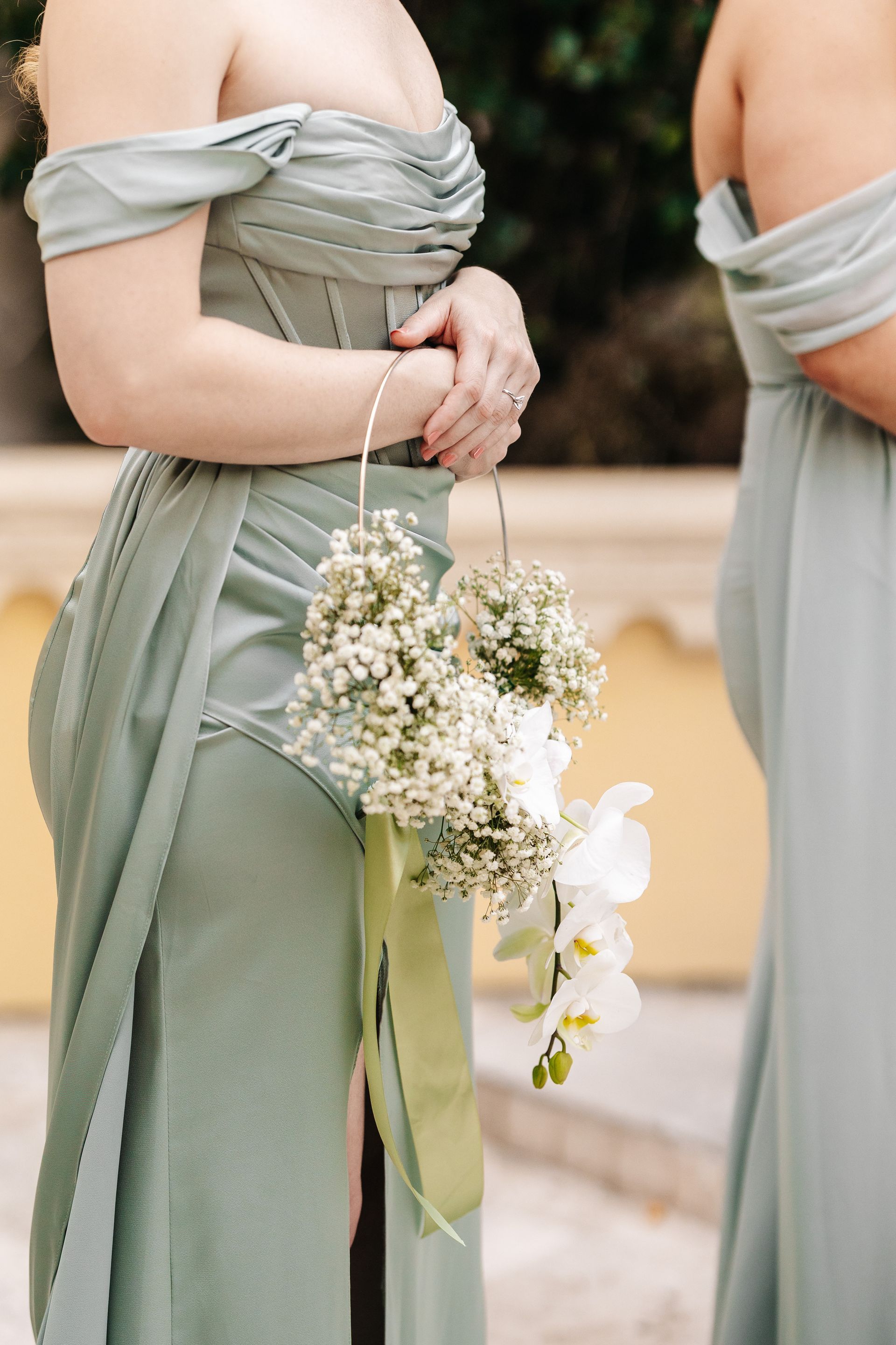 Two people in sage green off-shoulder dresses, one holding a floral basket.