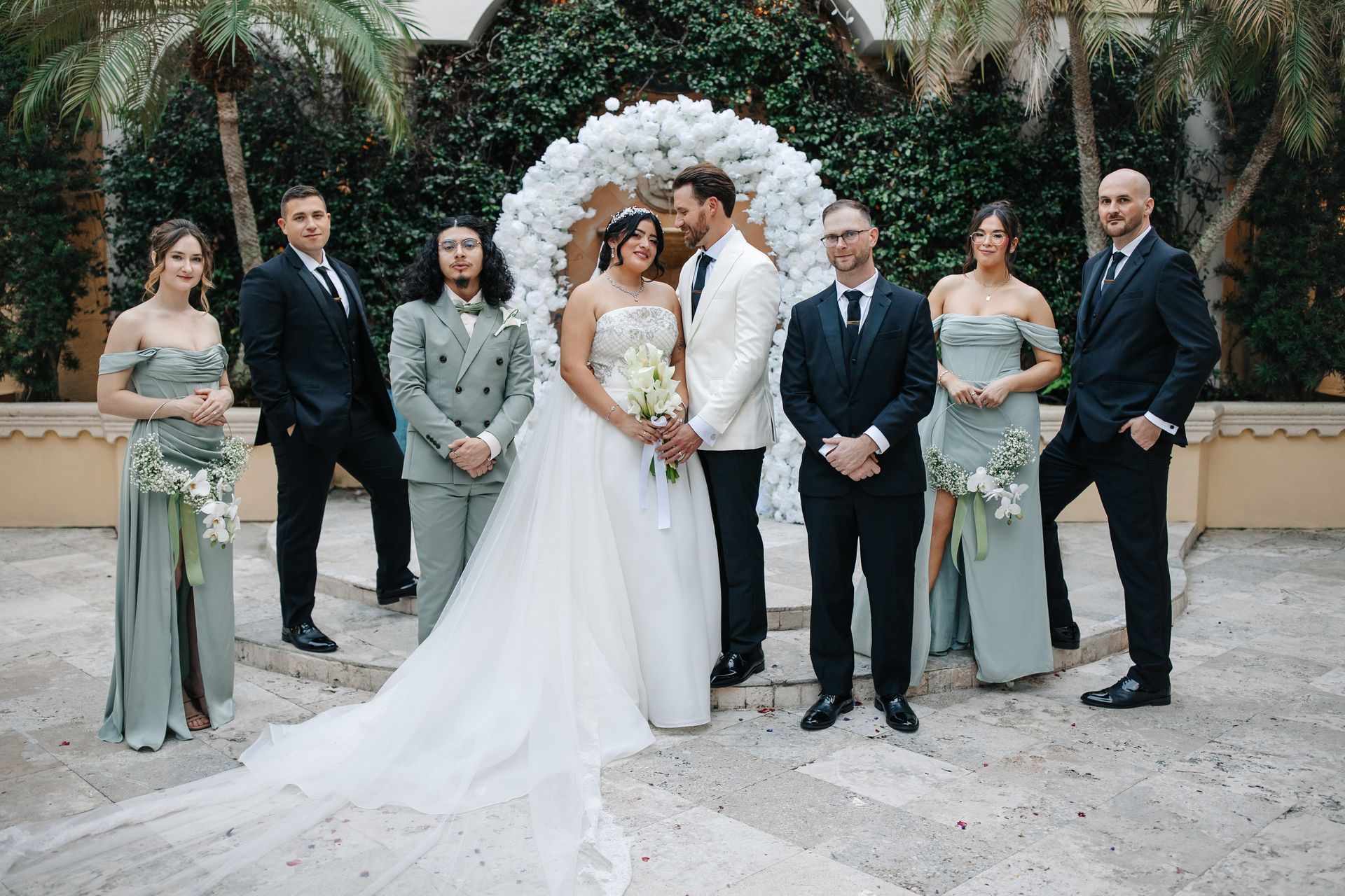 Wedding party posing in front of white floral arch. Bride in white dress, groom in white jacket. Others in black or sage.
