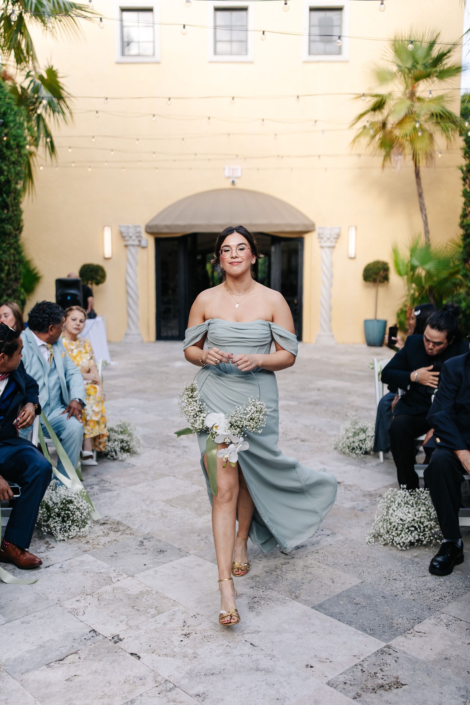 Woman in green dress walks toward camera, holding flowers at outdoor wedding.