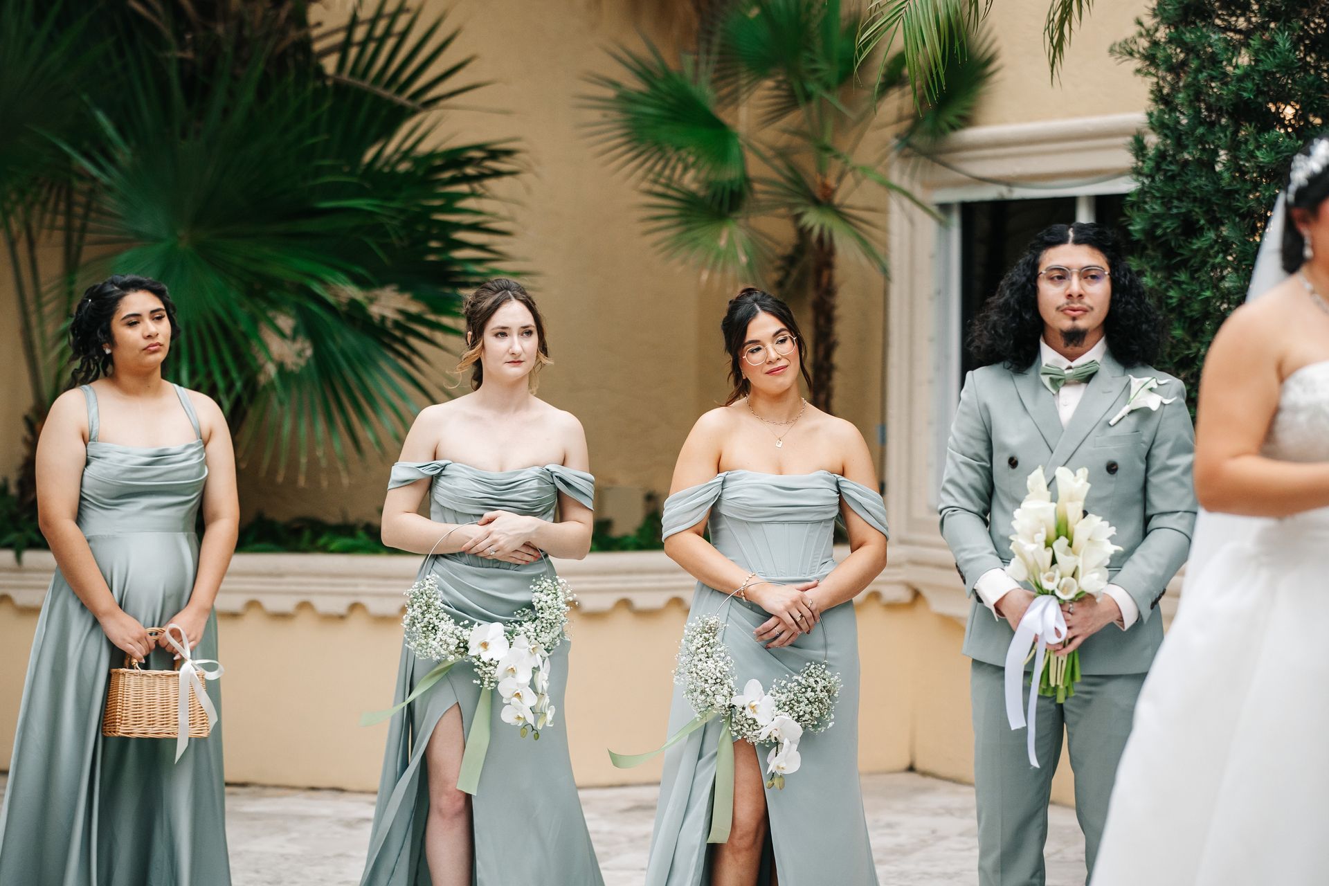 Wedding ceremony: Bride and bridesmaids in dusty green dresses stand with a groomsman holding flowers outdoors.