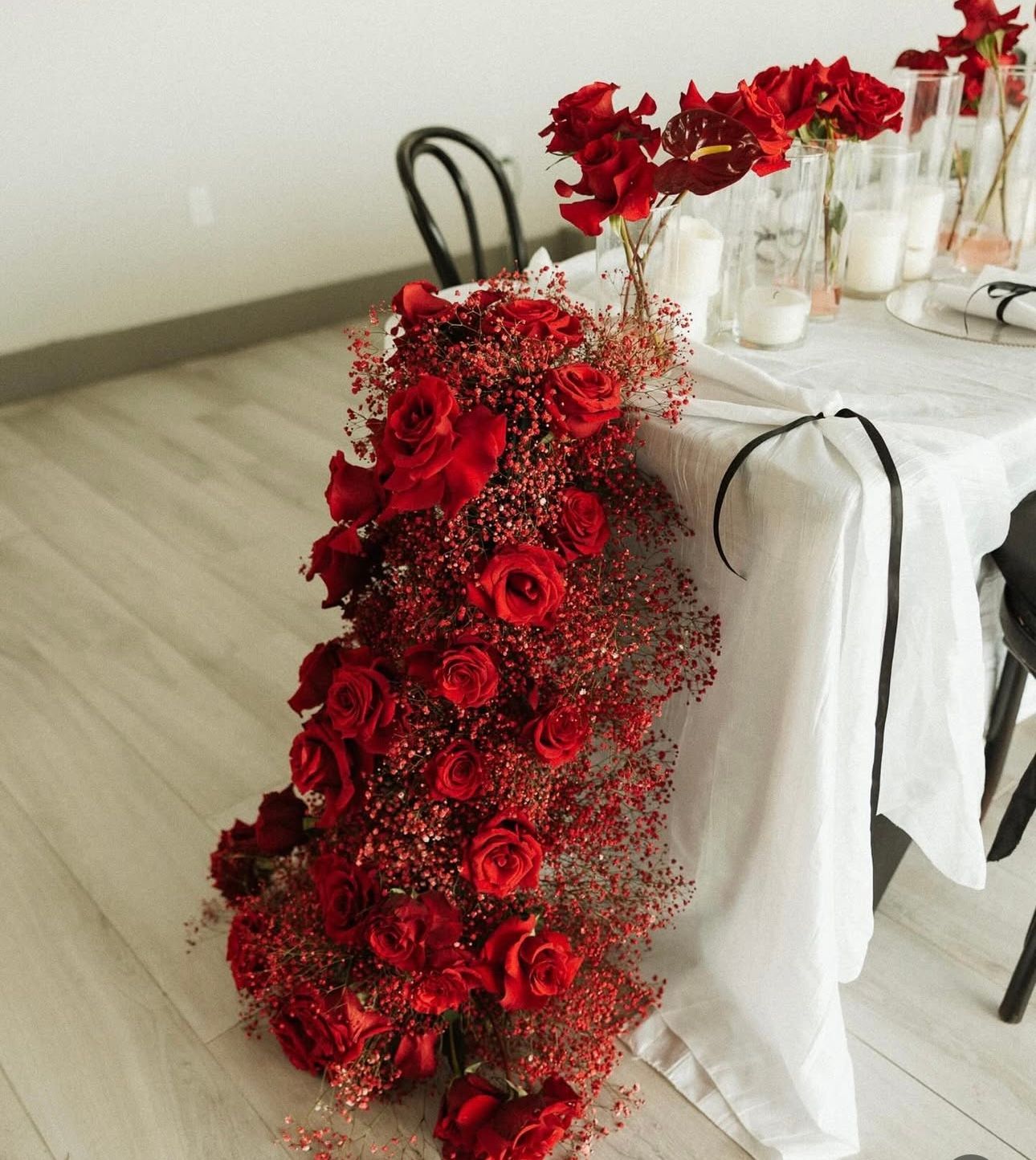 Red rose floral arrangement cascading down a white draped table with candles and chairs.