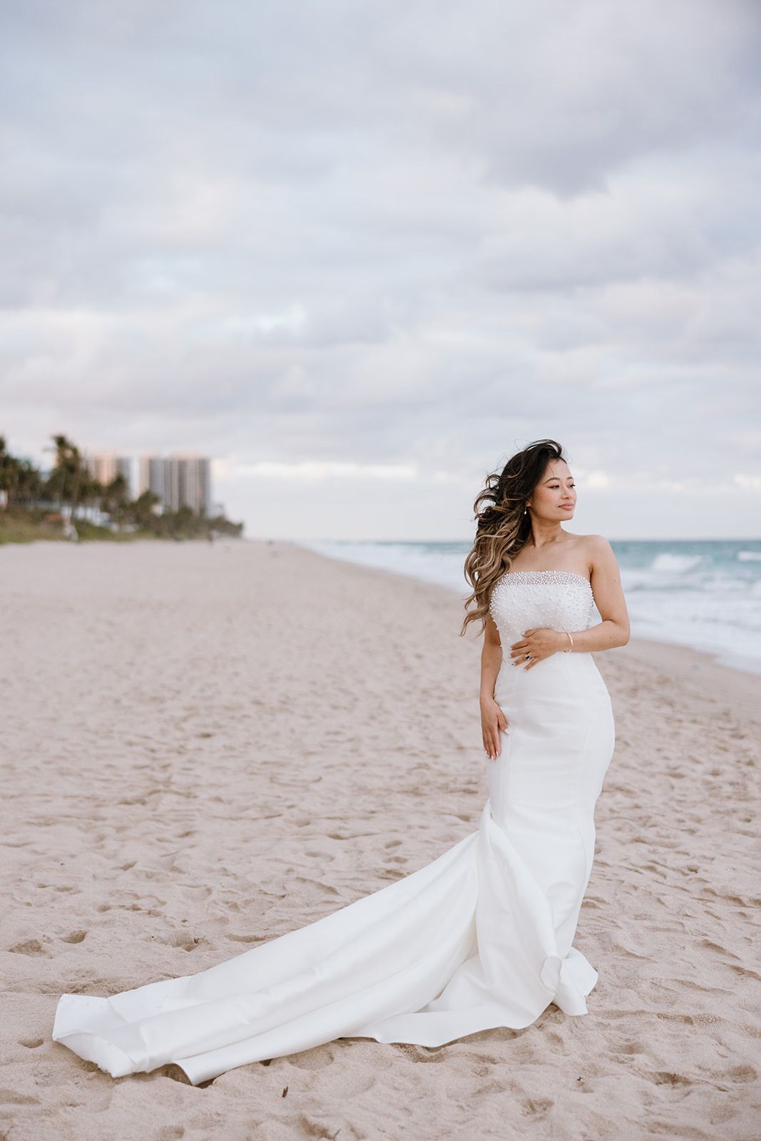 A pregnant woman in a wedding dress is standing on the beach.