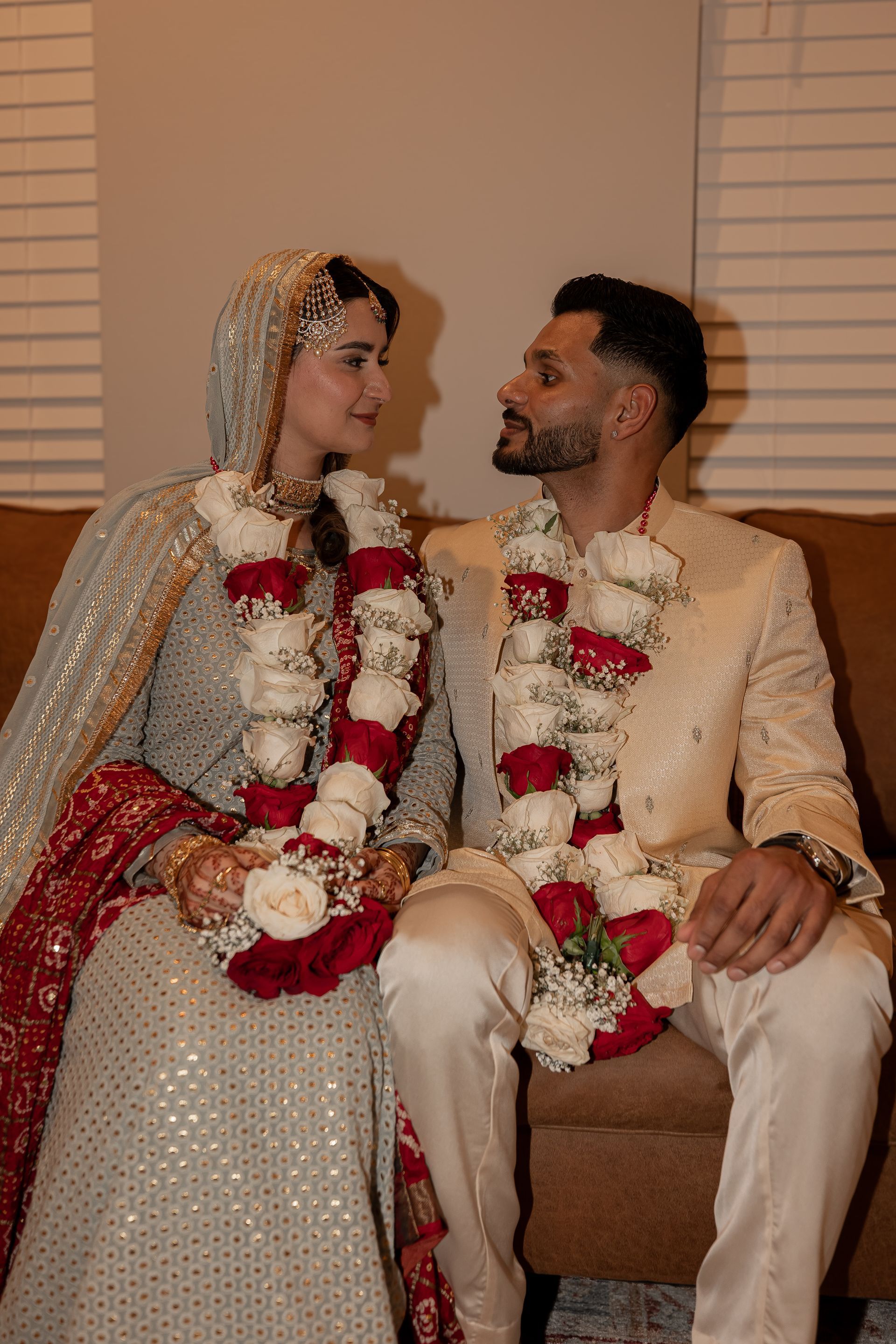 Couple in formal attire, seated, gazing at each other. They wear garlands. Beige and red color scheme.