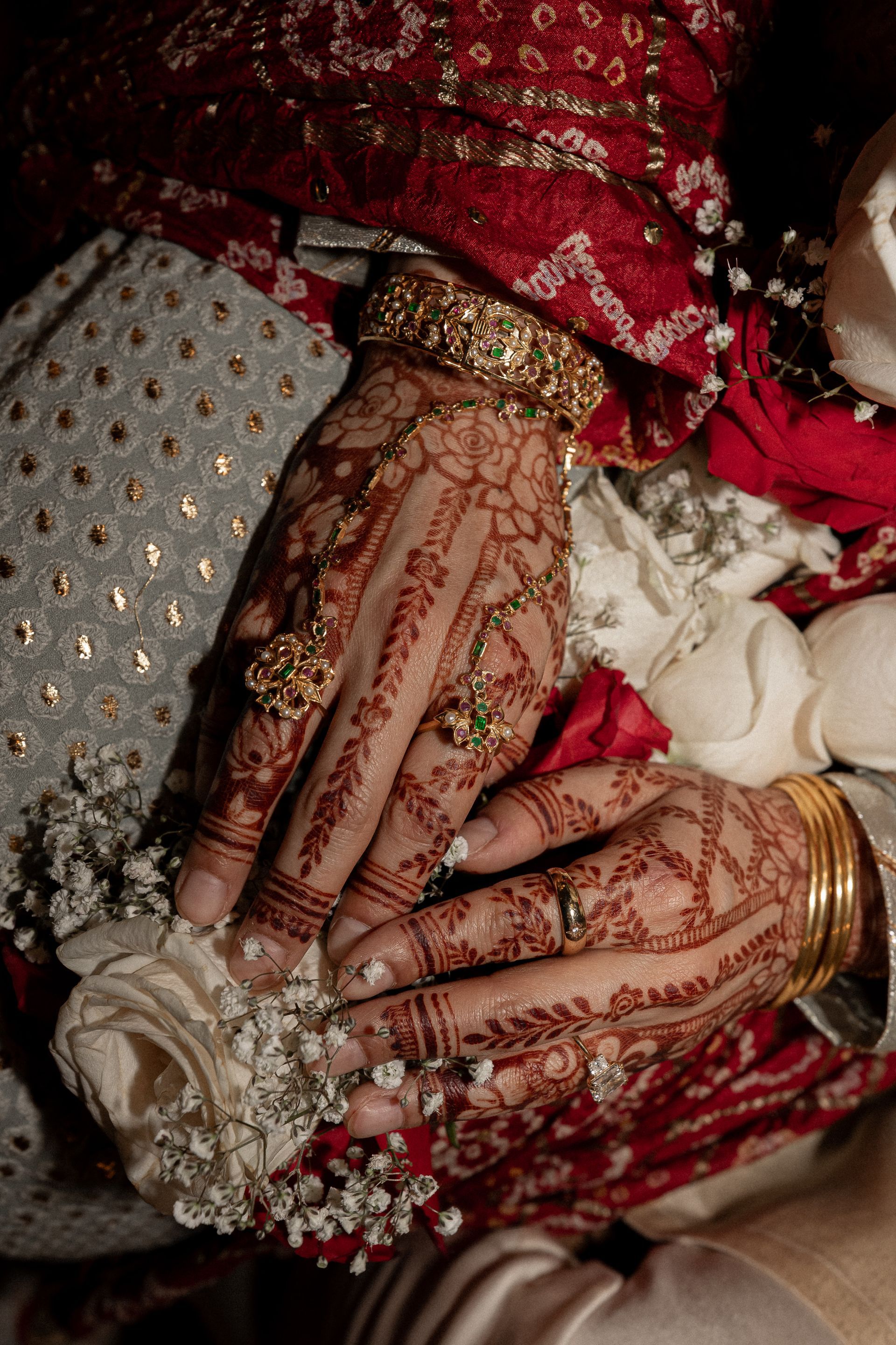 Hands adorned with henna and gold jewelry, holding white flowers, with red and white fabric in the background.