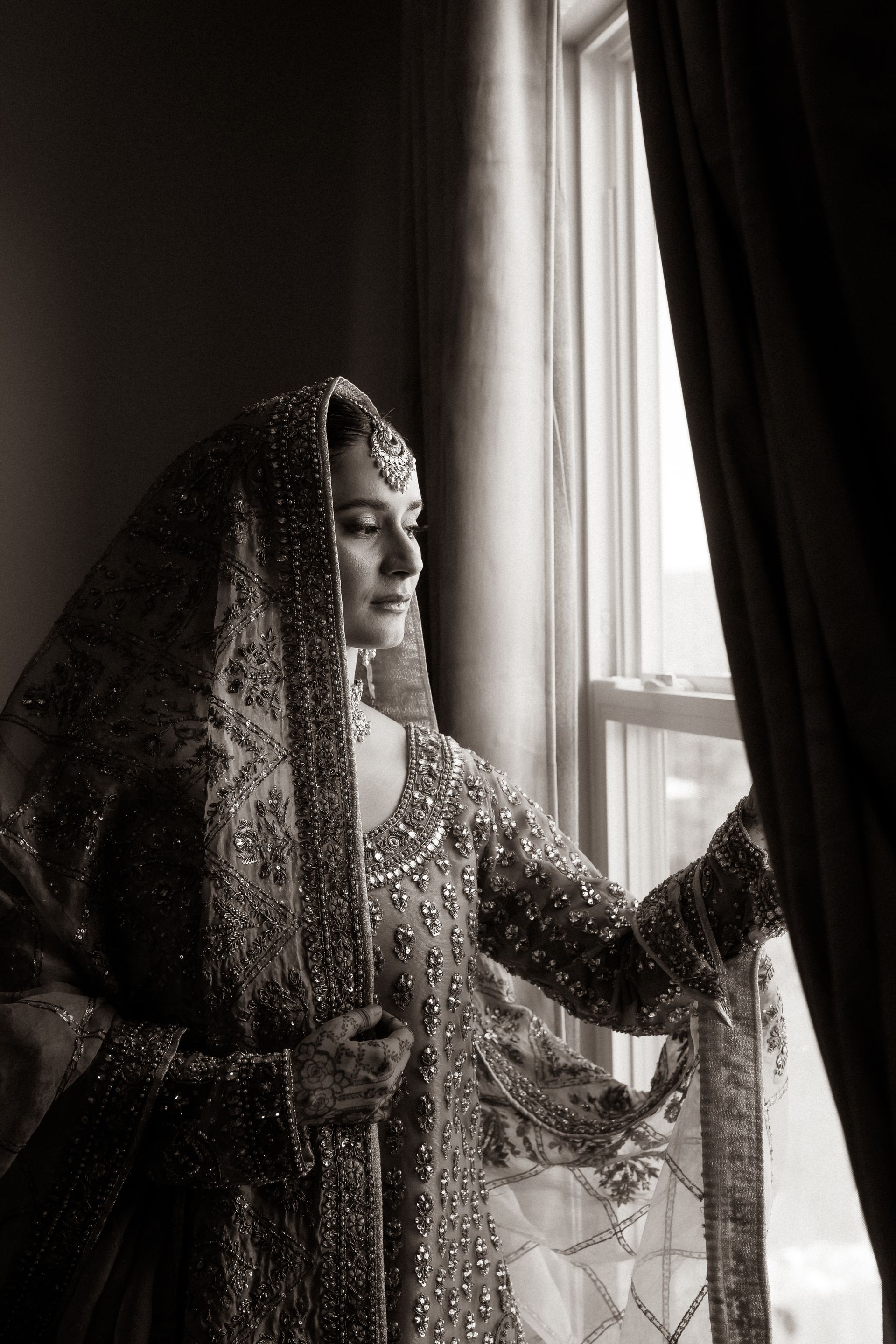 Woman in ornate, jeweled attire gazes out a window, holding the curtain. Black and white photo.