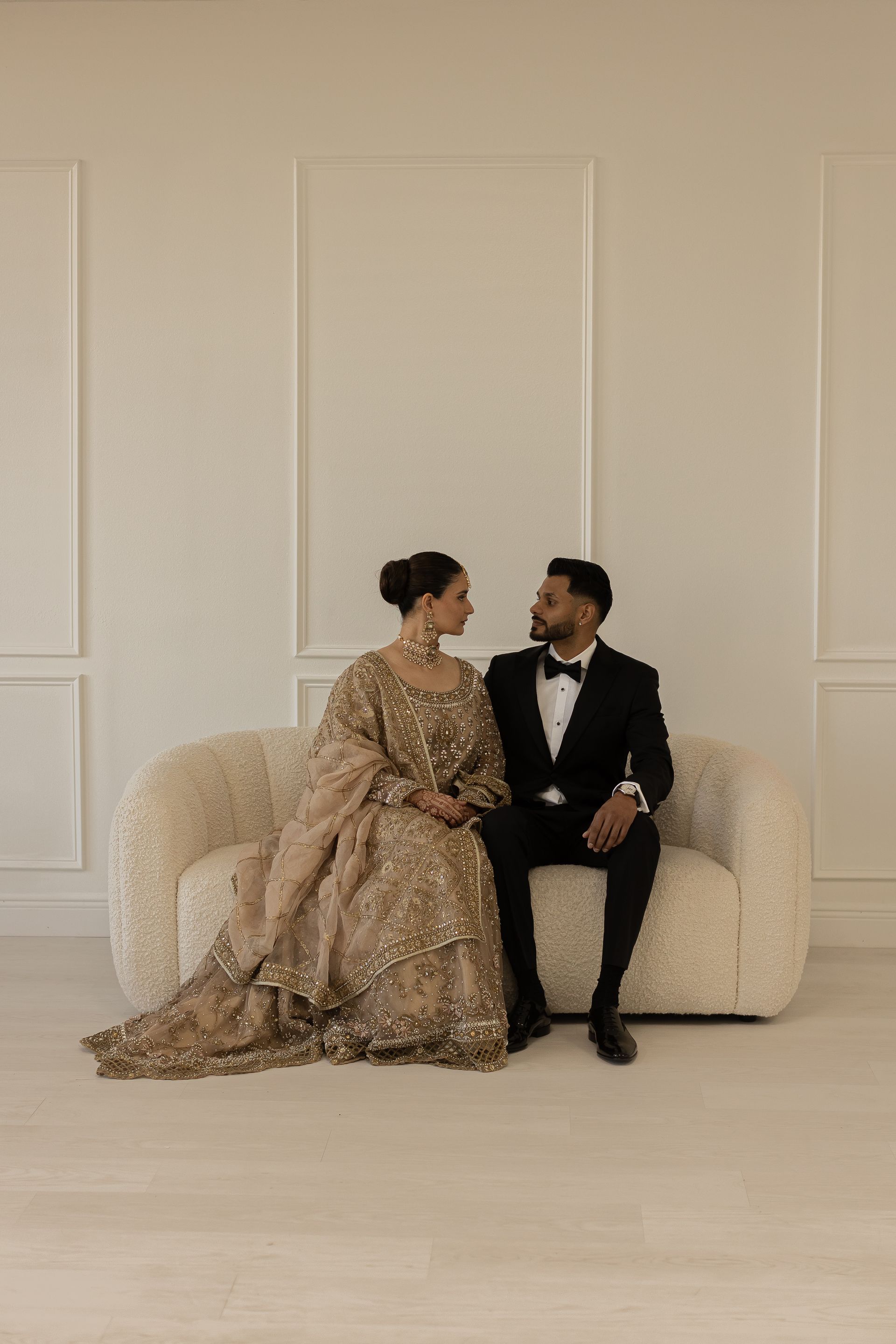 Couple in formal wear on a cream couch, looking at each other. Beige walls.