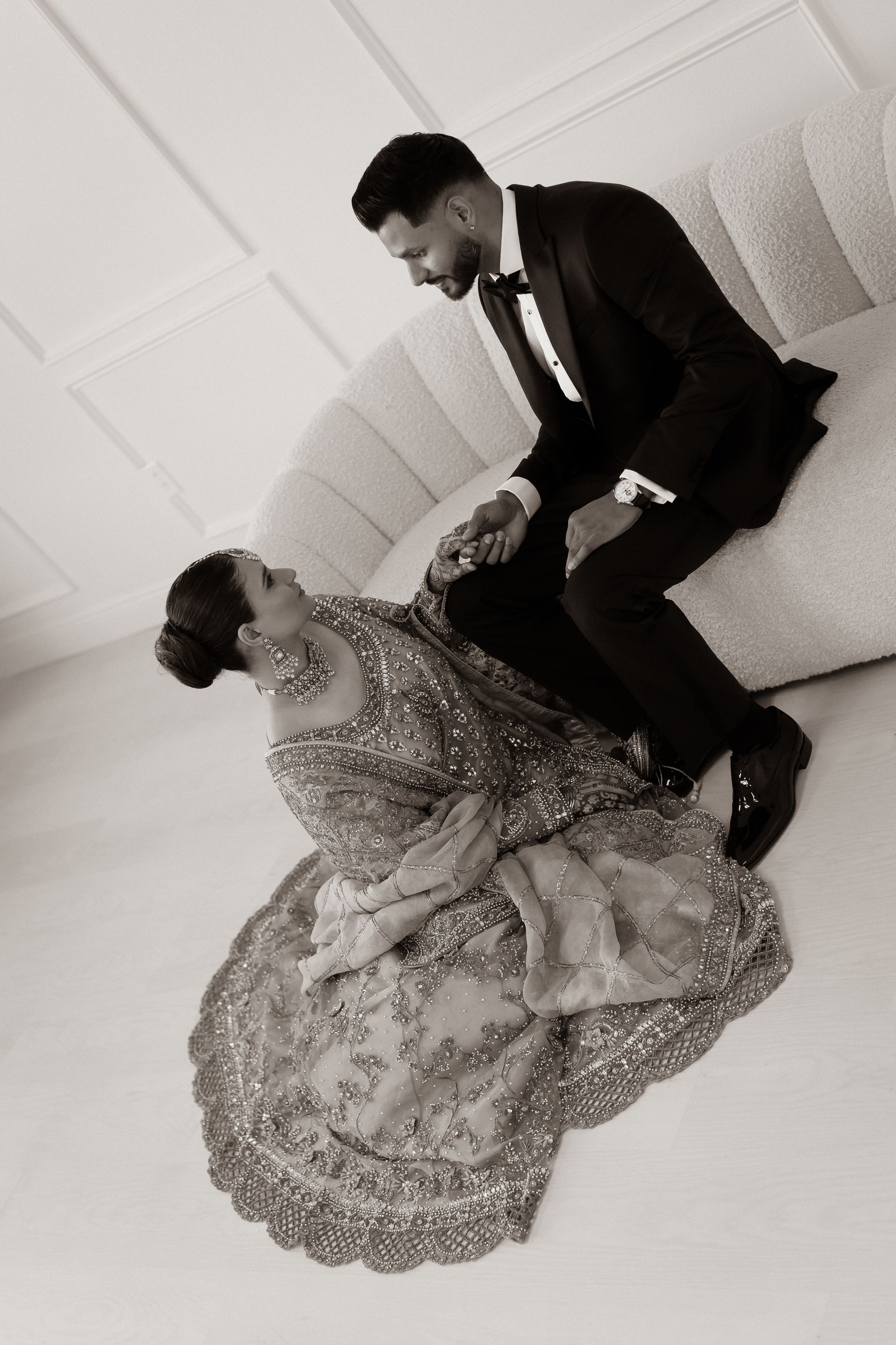 Couple in formal attire, posing on a curved sofa, looking at each other. Black and white photo.