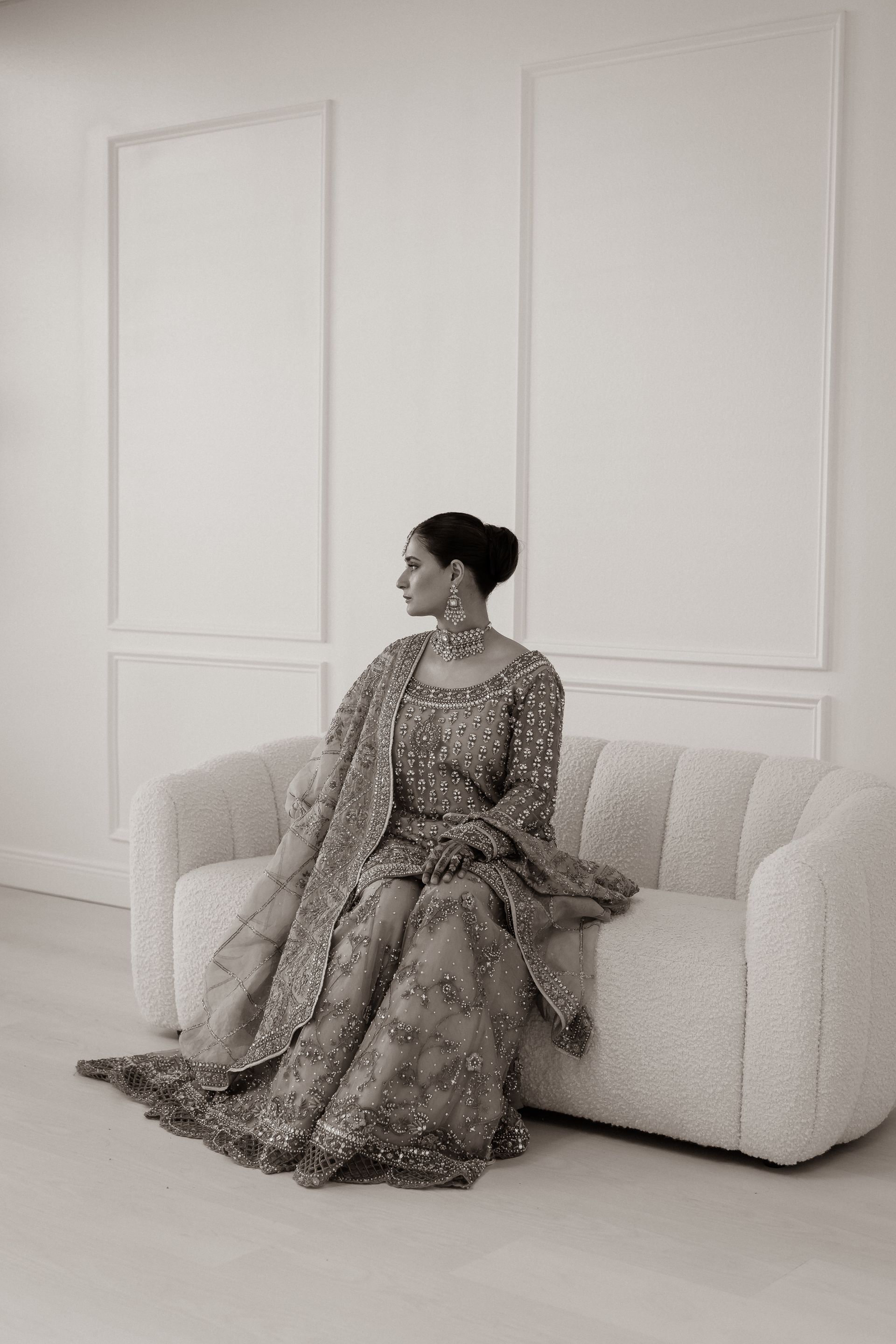 Woman in embellished gown sits on a cream-colored couch, looking to the side, against a white wall.