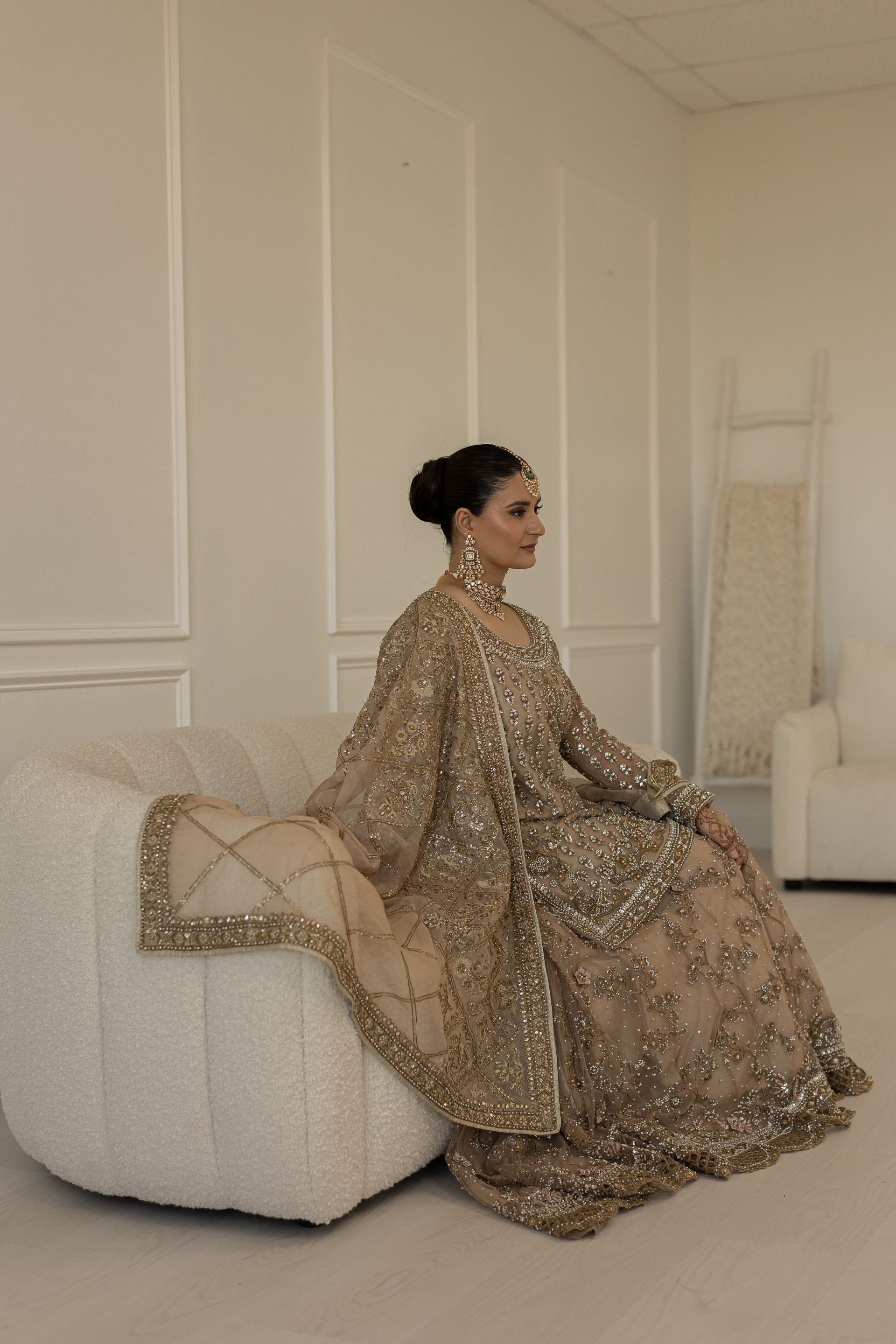Woman in gold embellished gown sits on a white ottoman, holding a matching shawl.