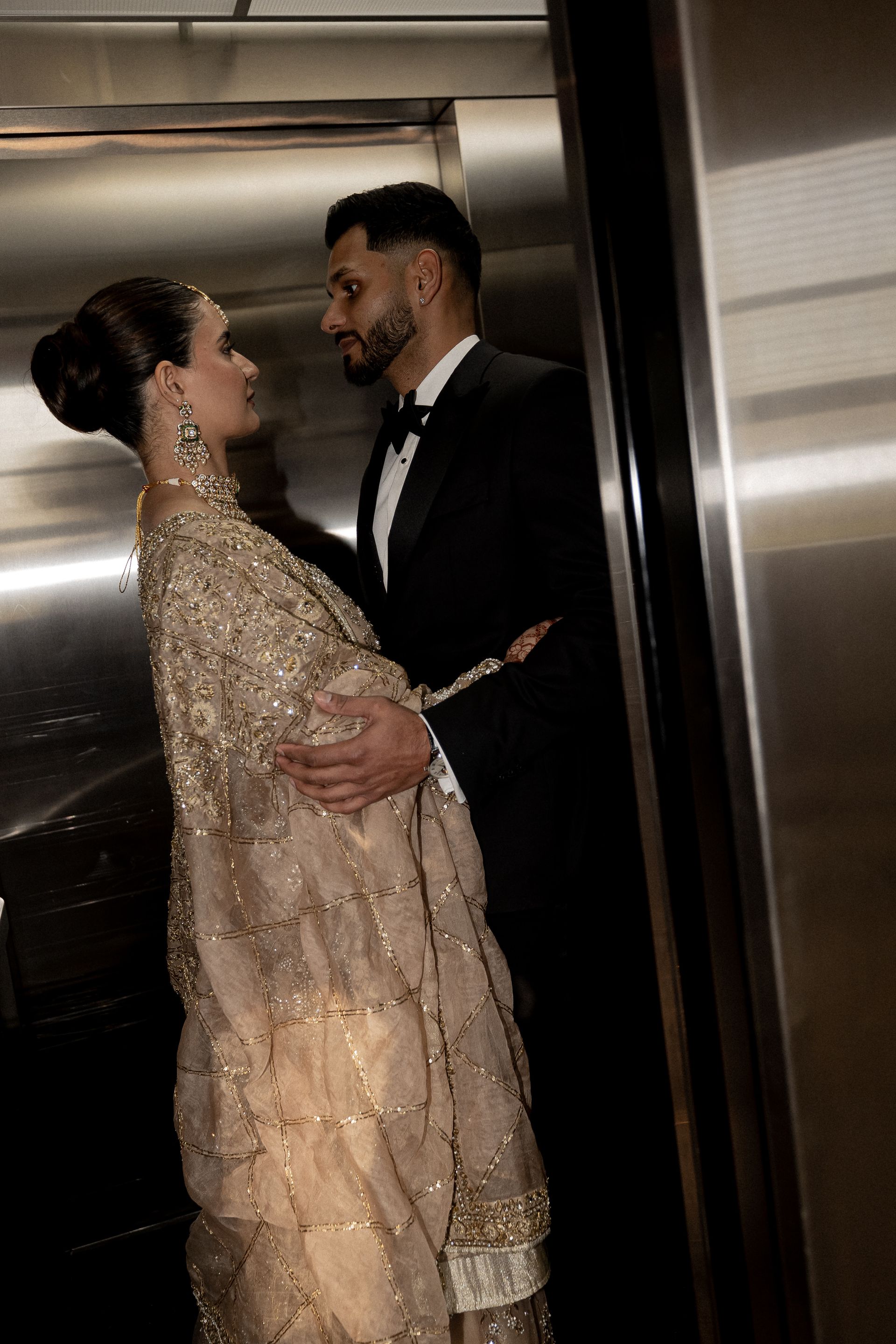 Couple in formal attire embracing in an elevator. Gold beaded gown, black tuxedo.