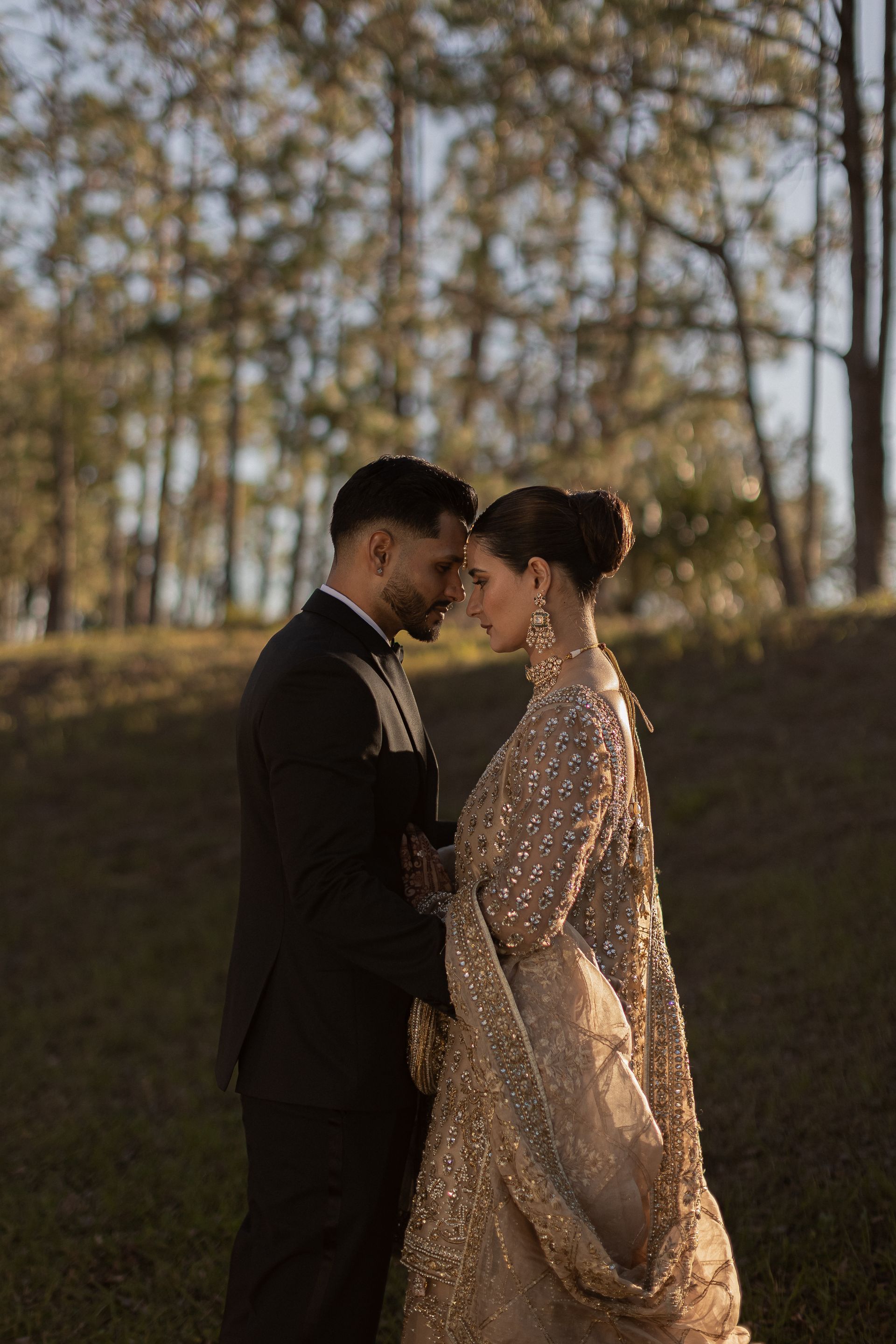 Couple in formal attire, forehead to forehead, golden light, outdoor setting.