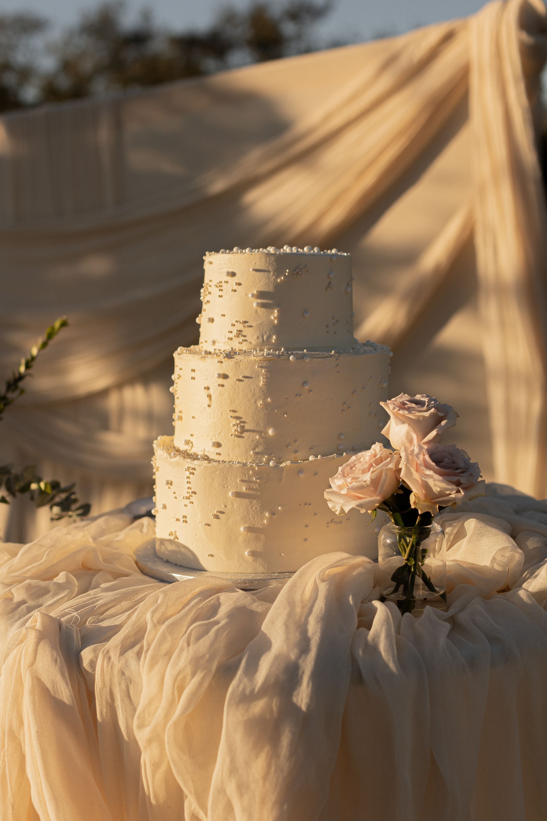 Three-tiered white wedding cake with silver sprinkles and flowers on a draped table outdoors.