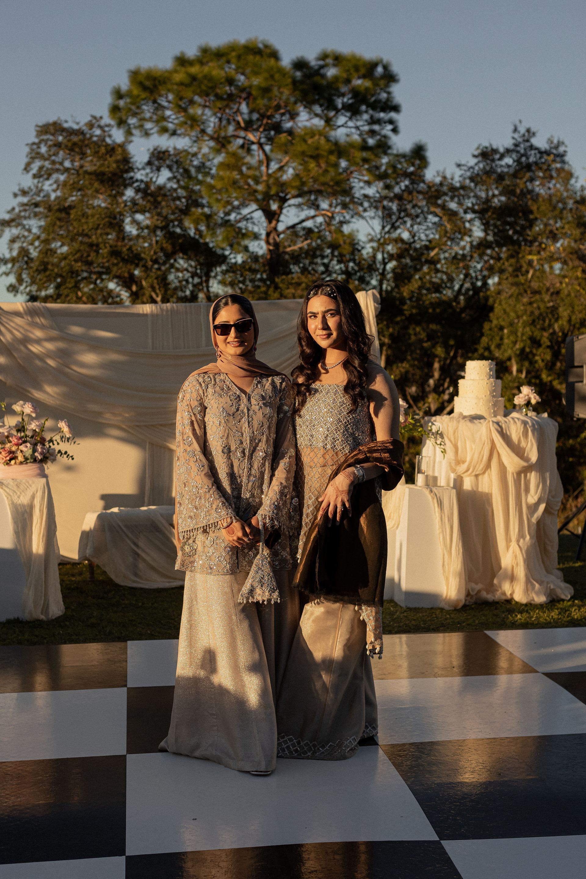 Two women in embellished gowns pose on a checkered floor outdoors, a wedding cake in the background.