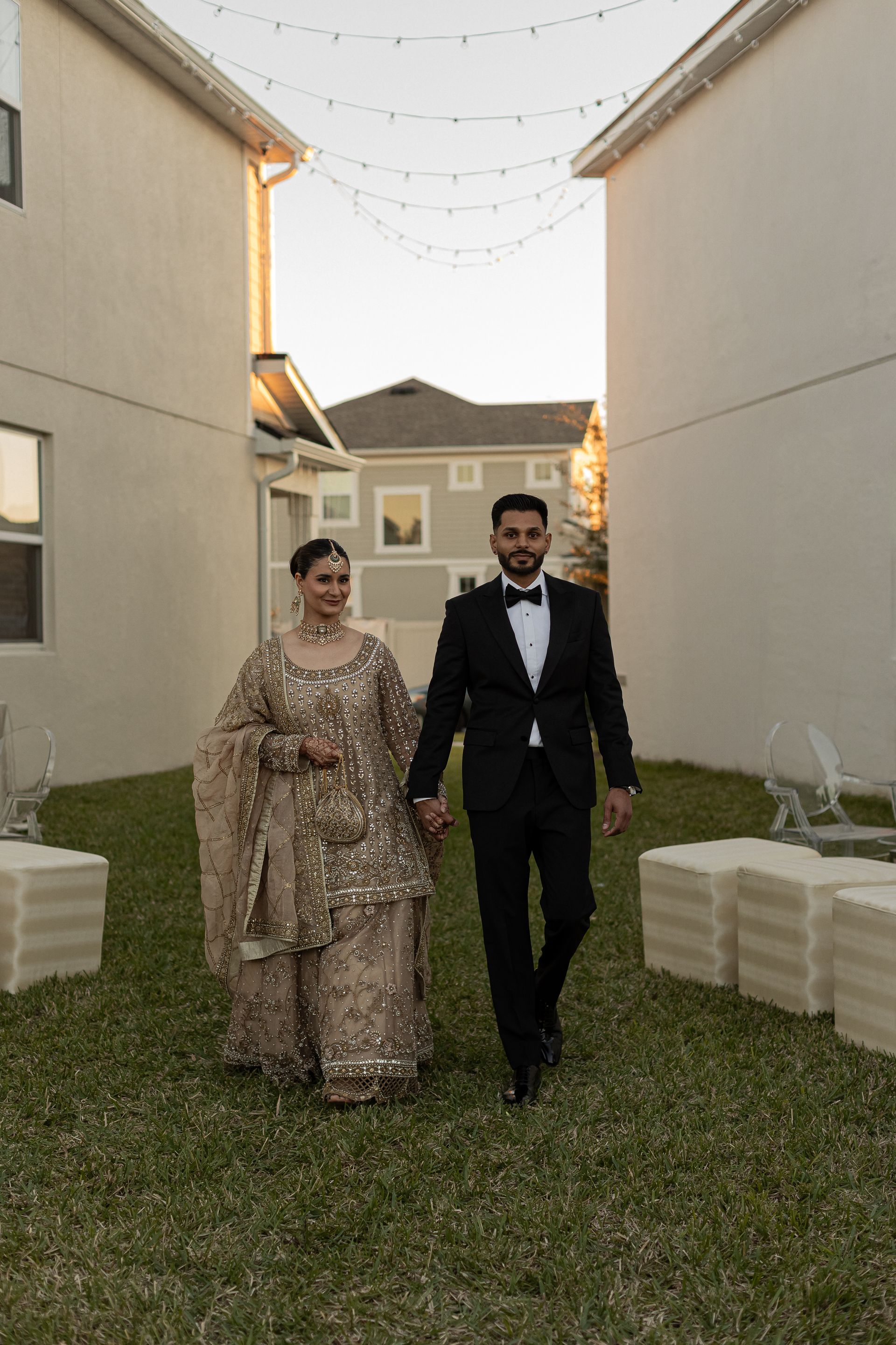 Couple in formal attire walking, holding hands, in a backyard setting, under string lights.