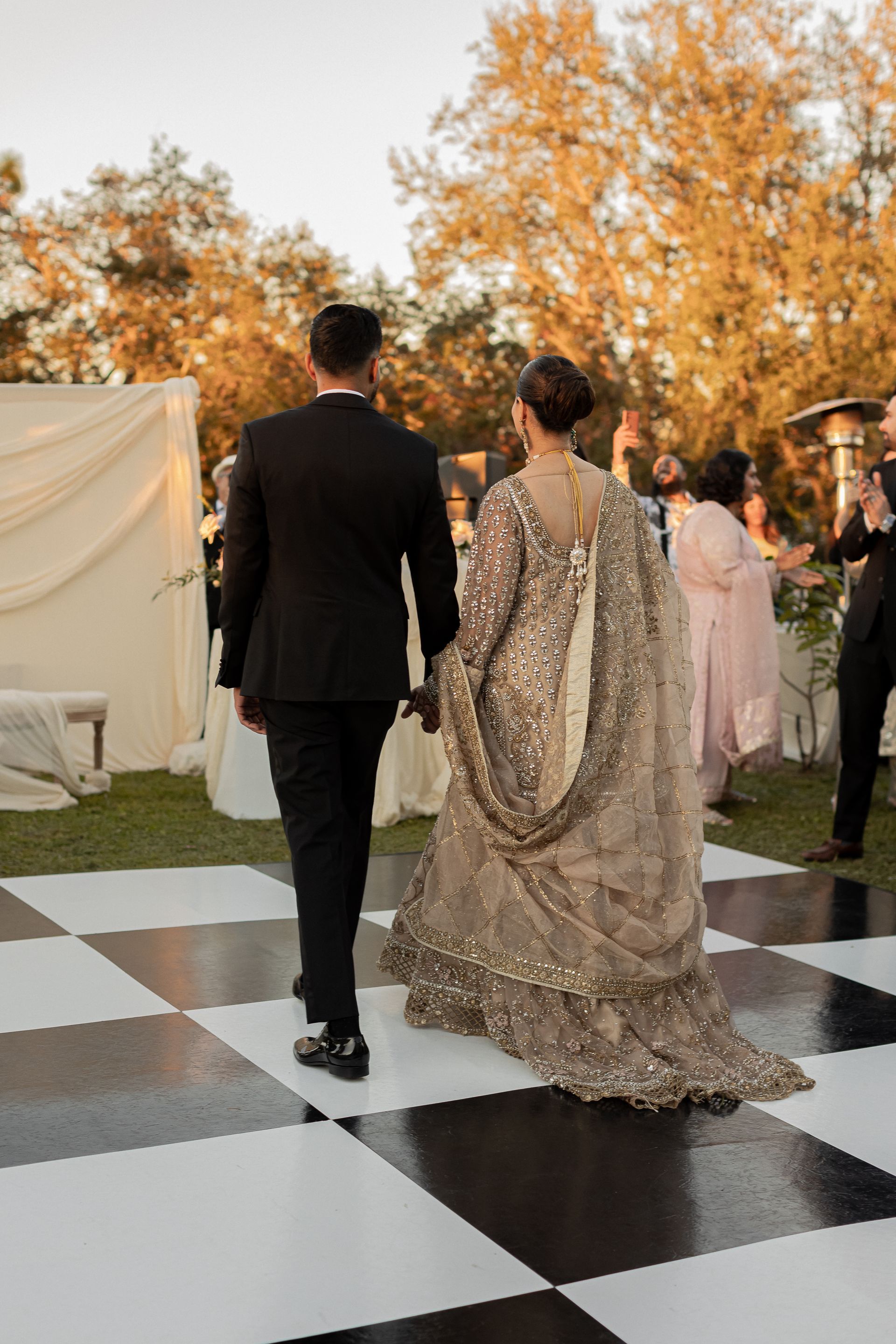 Couple walking on checkered dance floor. The woman wears a gold embellished gown with a long train. The man wears a black suit.