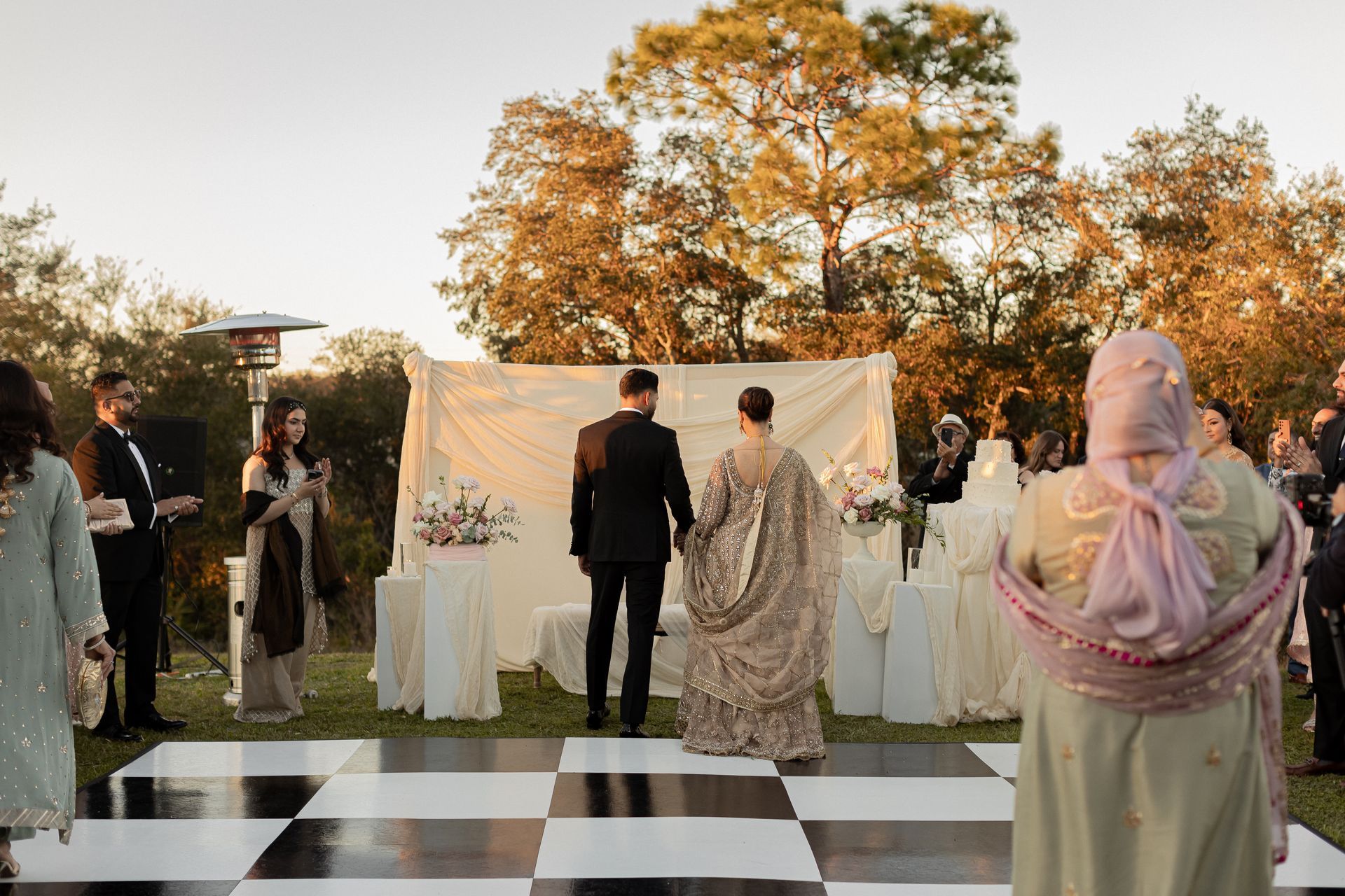 Couple holding hands on a checkered dance floor during an outdoor wedding ceremony; guests watching.