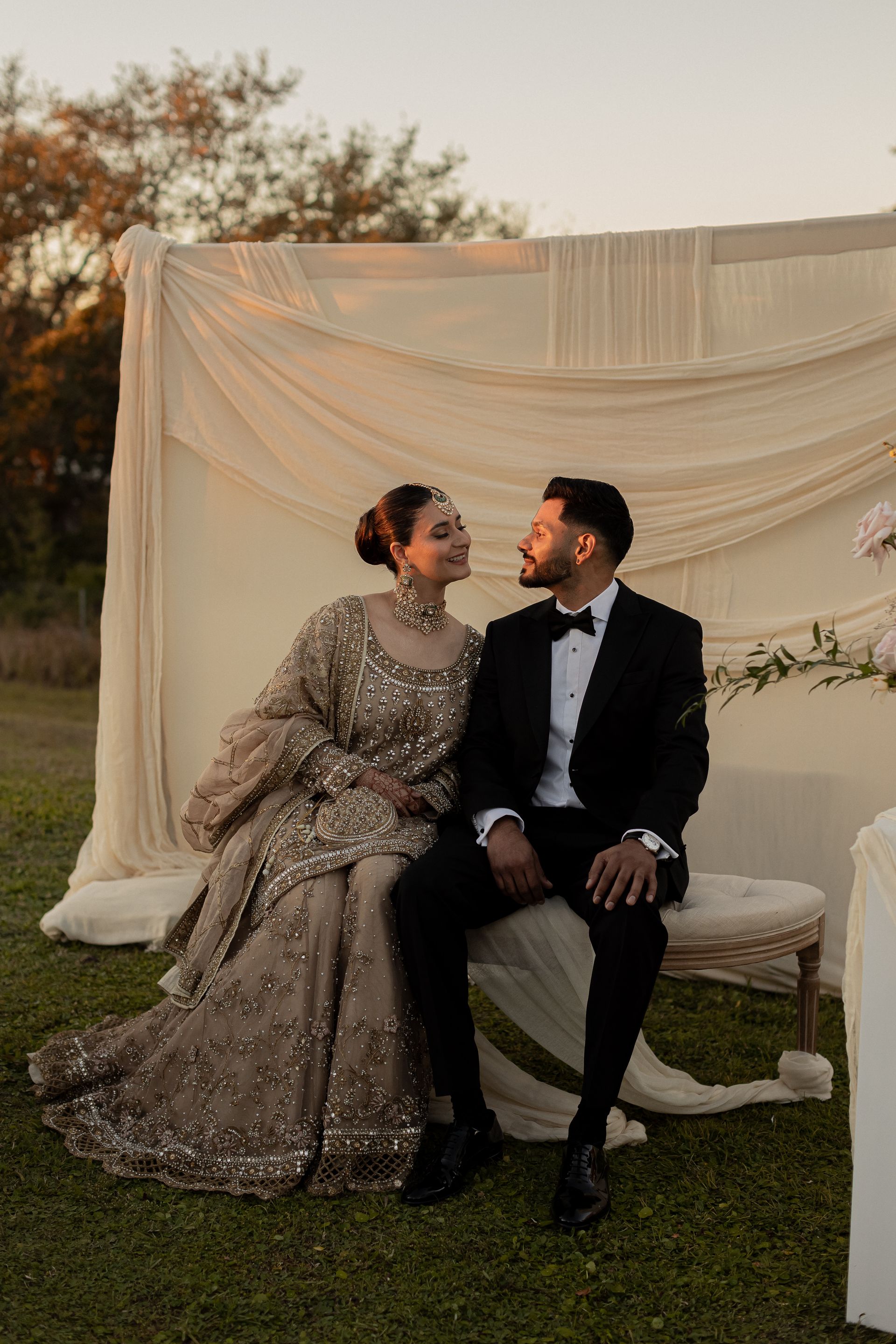 Couple in formal attire seated outdoors, looking at each other. Beige backdrop, sunset lighting.