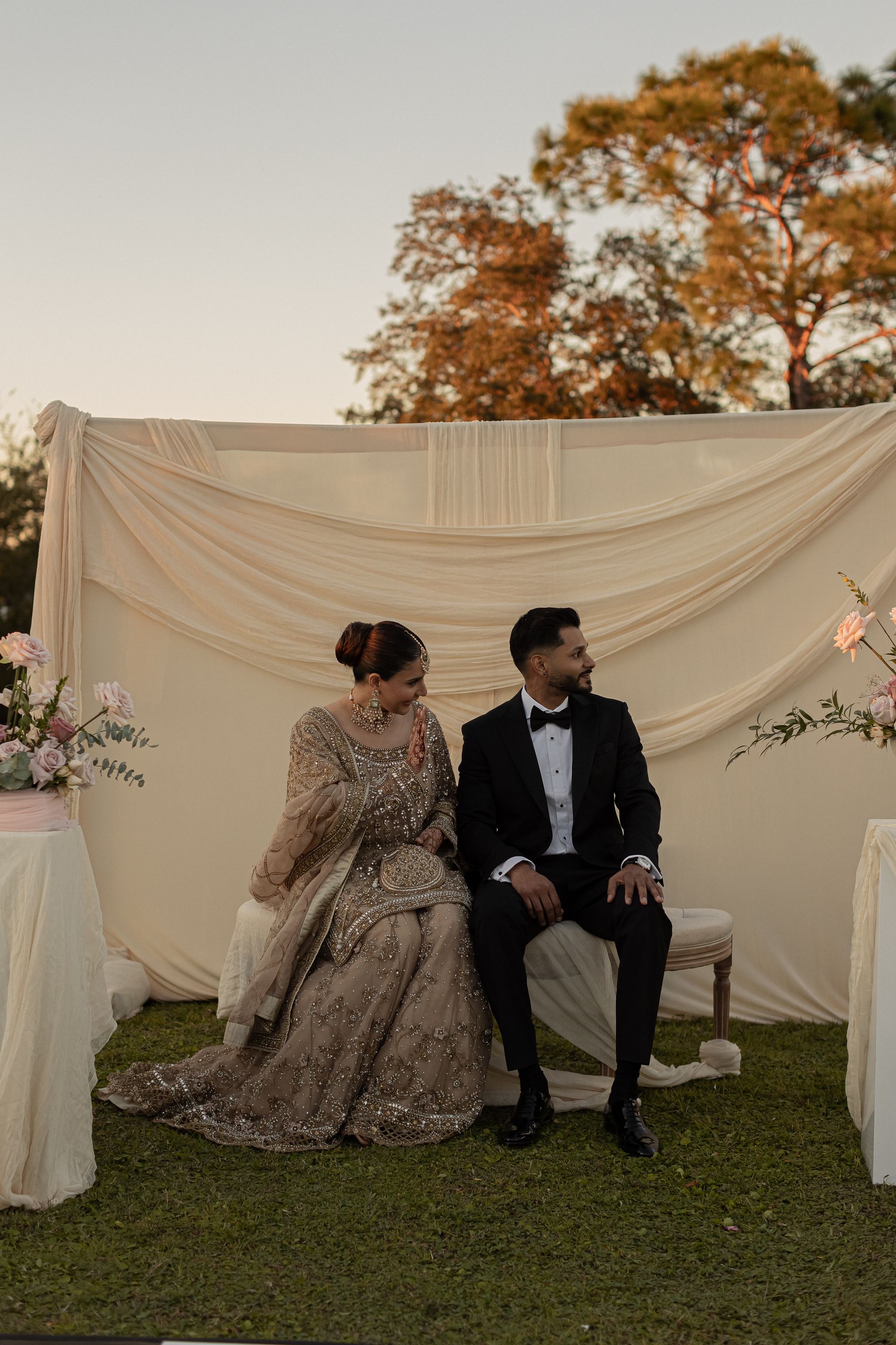 Couple in formal attire seated outdoors, looking away. Beige backdrop, sunset.