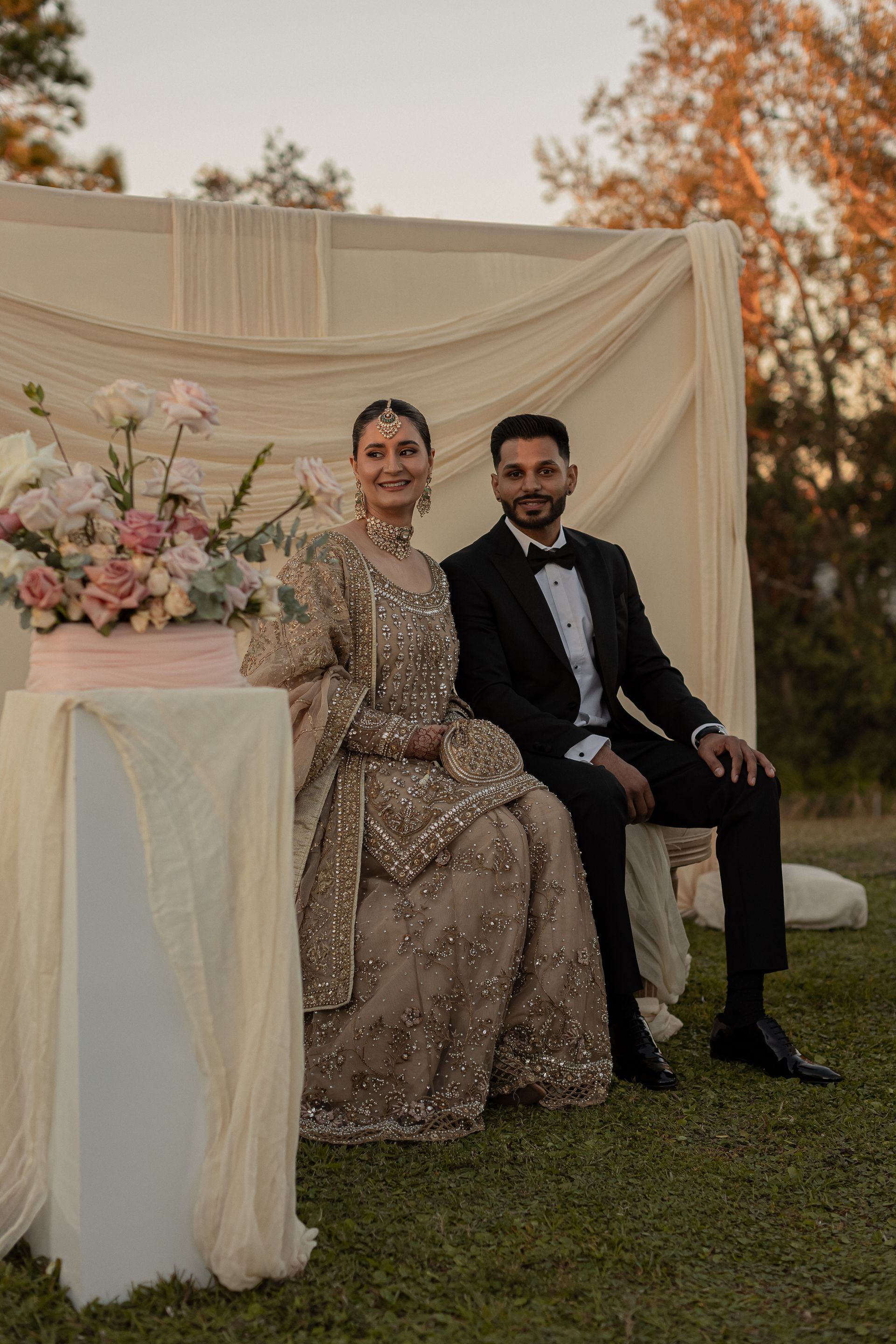 Couple in wedding attire seated outdoors near a floral display. Beige and gold clothing, black suit, bow tie, flowers, greenery.
