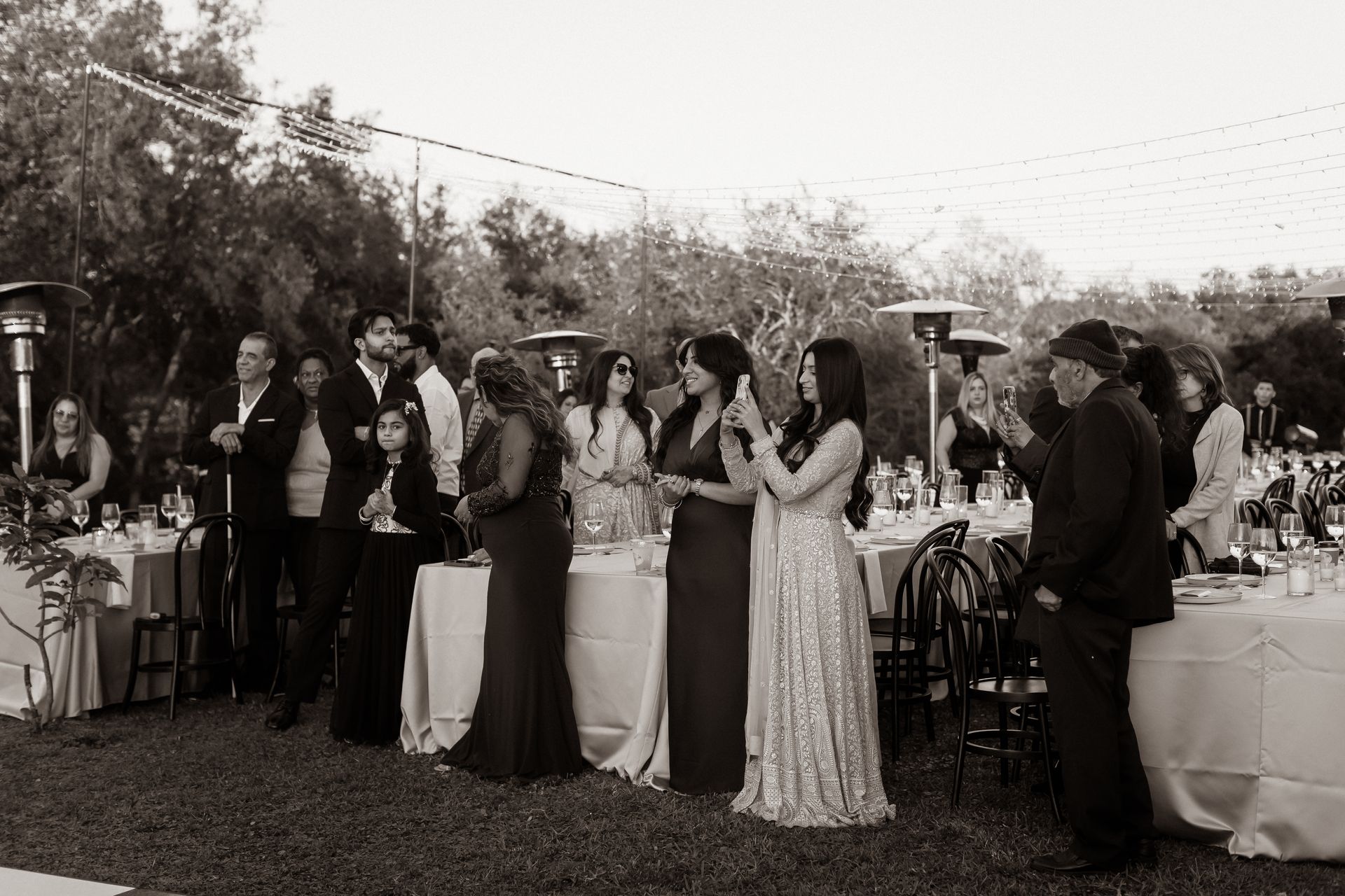 Guests at outdoor reception, tables set. Some people are talking, wearing formal attire.